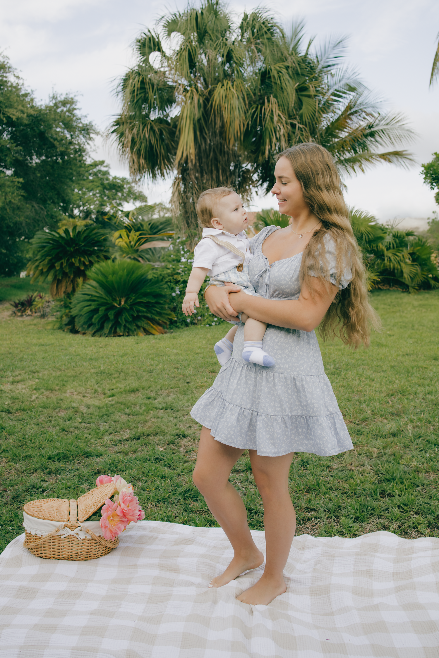 Mother and daughter in matching gray dresses dance together on a picnic blanket in a tropical garden setting.