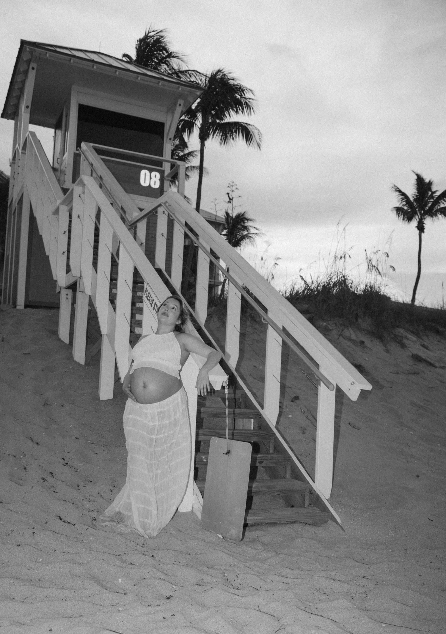 Black and white photo of person posing by beachside stairs with palm trees.