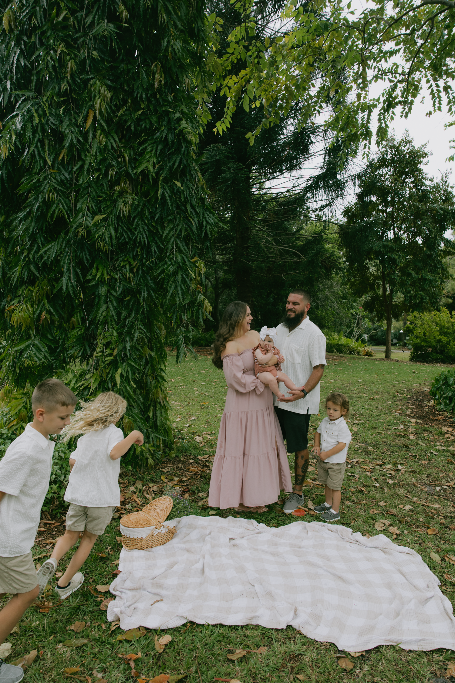Family having picnic under willow tree with white blanket spread on grass.