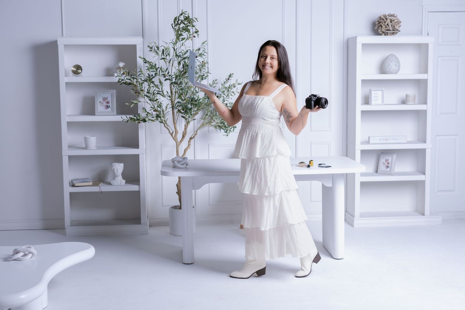 Woman in white tiered dress holding camera poses in bright white studio with shelves and indoor plant.