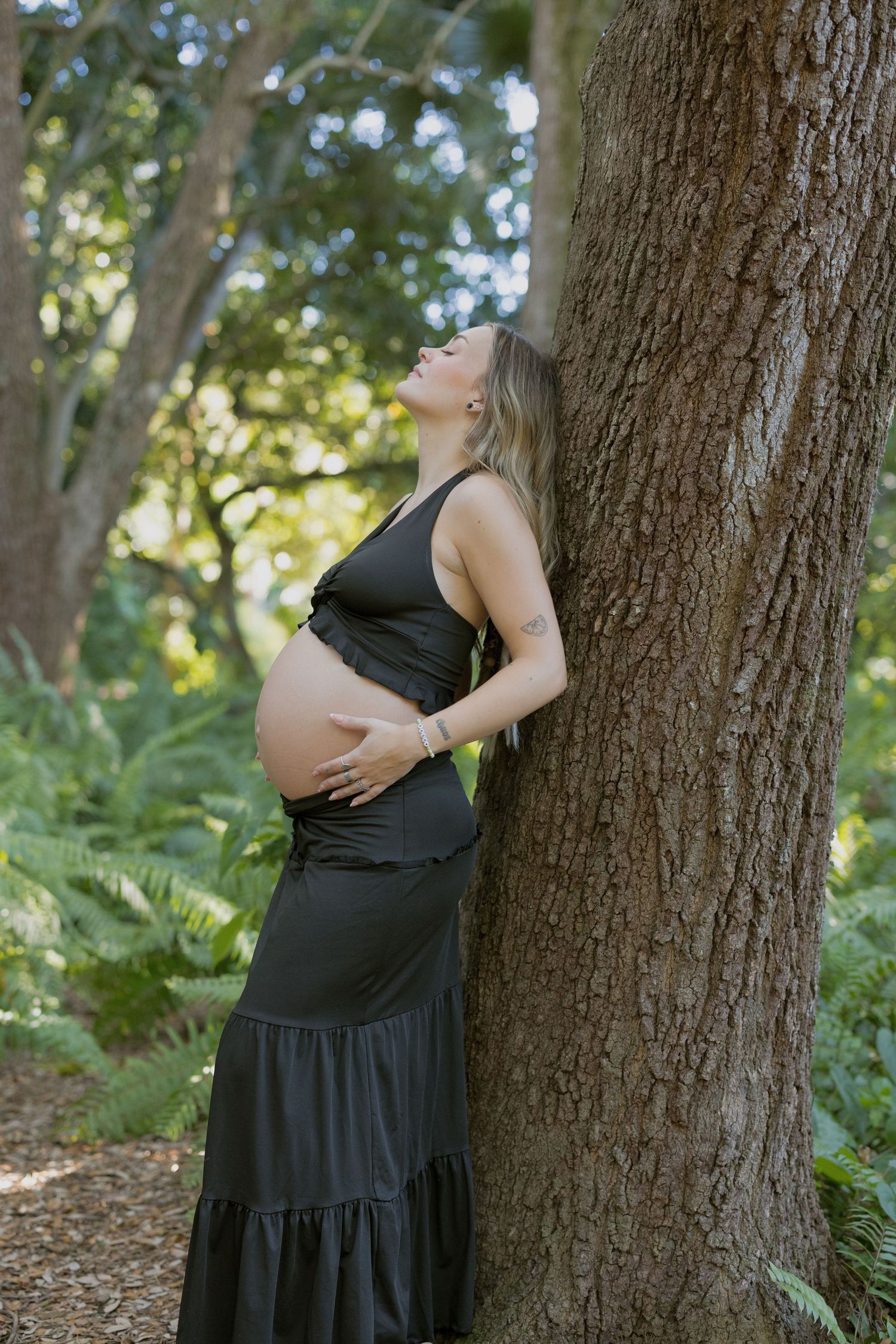 A person in a black dress poses against a tree trunk in a sunlit forest setting with green foliage in the background.