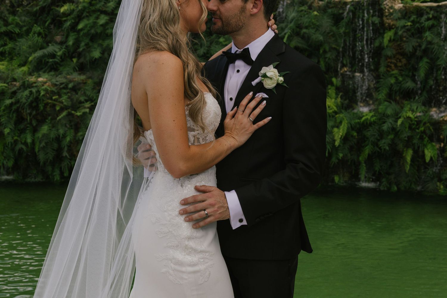 A couple shares a romantic kiss during their outdoor wedding ceremony surrounded by lush green gardens.