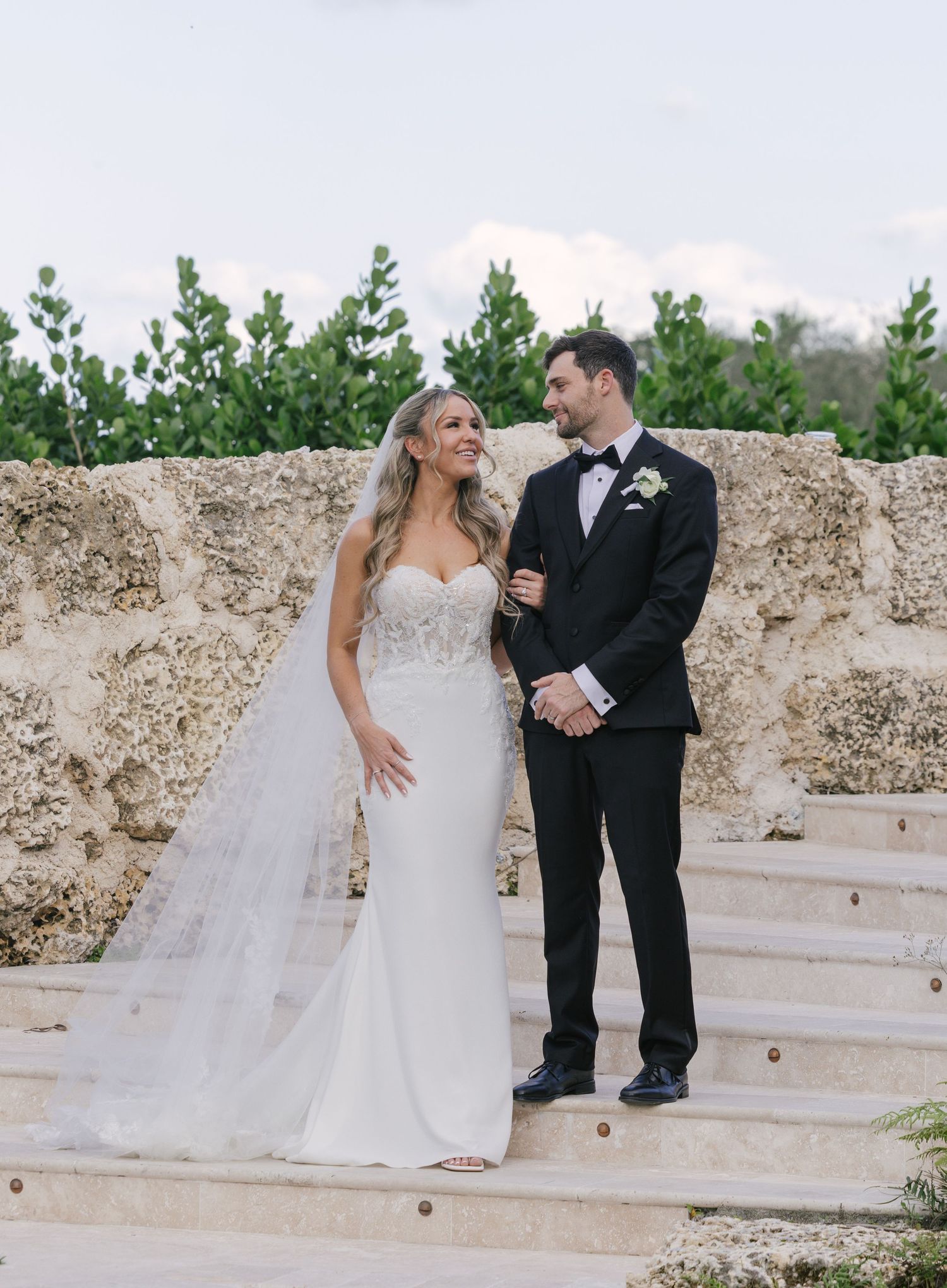 A romantic wedding couple shares intimate moments on stone steps in a garden setting with lush greenery in the background.