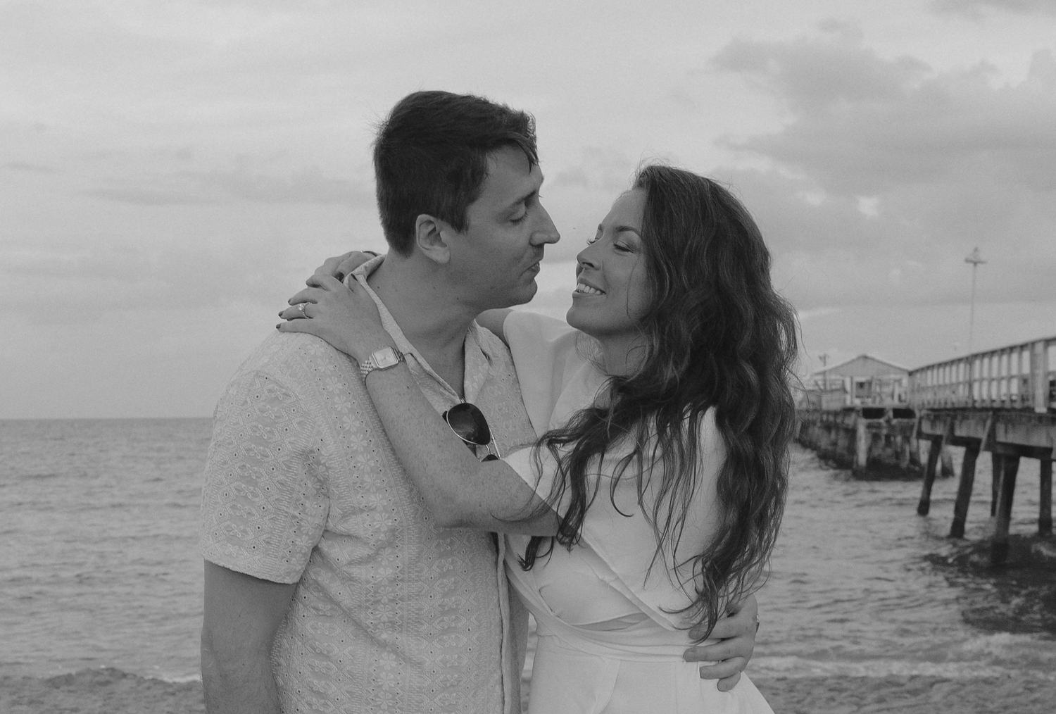 A romantic black and white photo series showing a couple sharing tender kisses by a pier at the ocean.