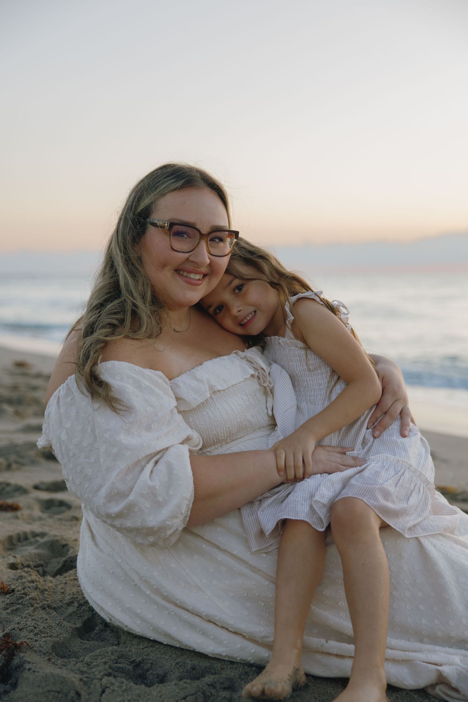 Two people wearing white share an embrace while sitting on the beach at sunset.