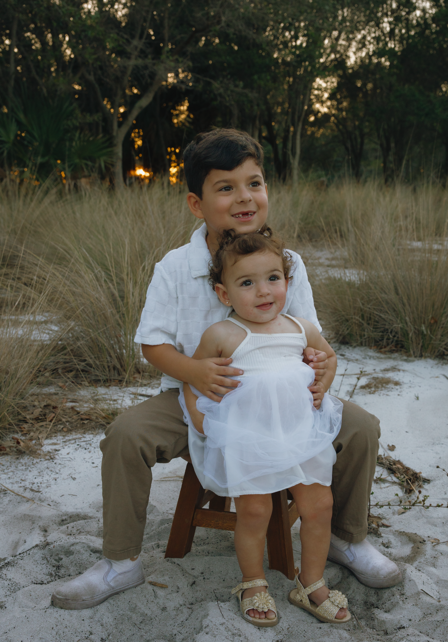 Two children share playful moments while sitting together on sandy ground with tall grass nearby.