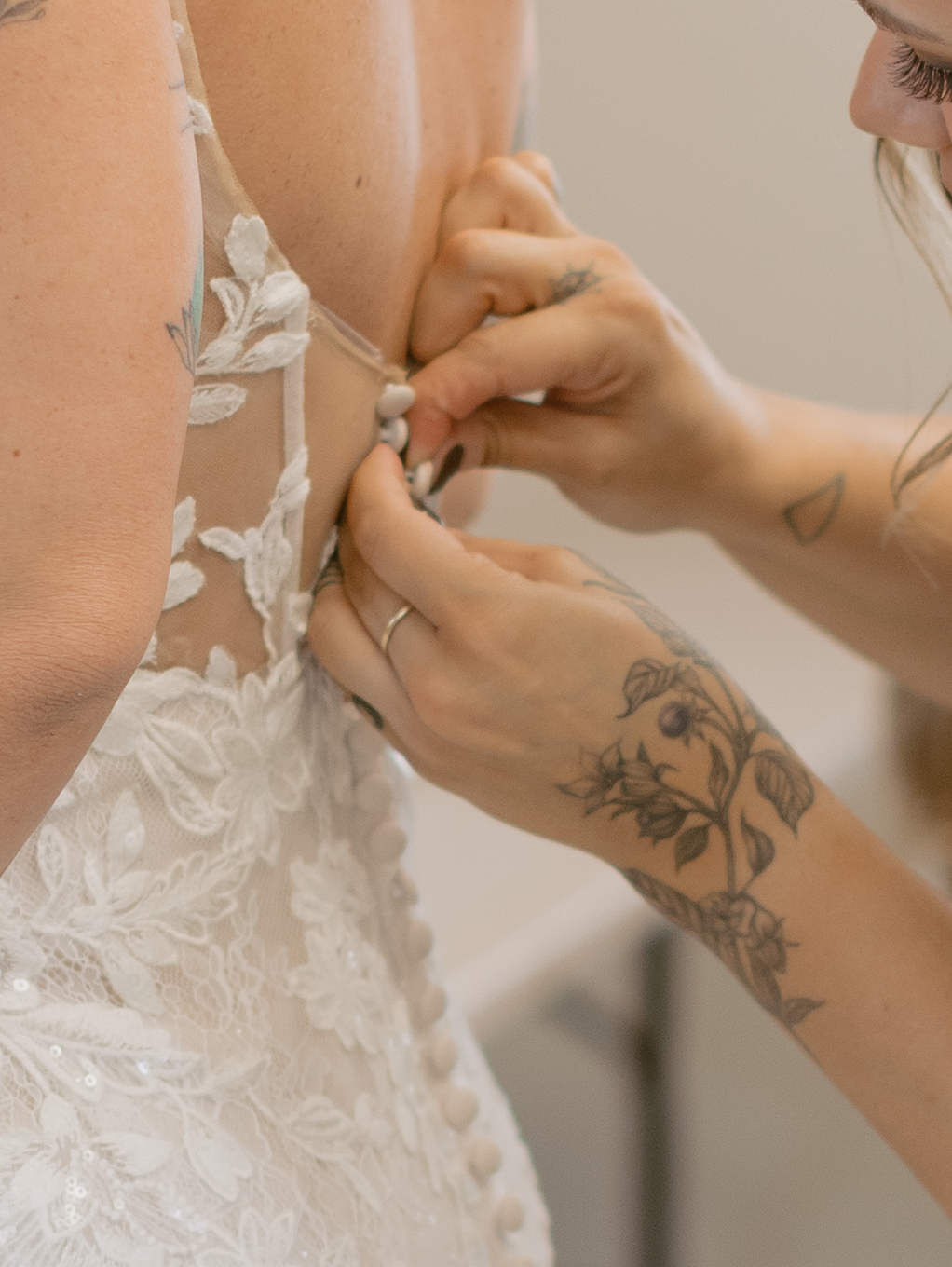 Hands with floral tattoos button up the back of an intricate white lace wedding dress in a delicate moment.
