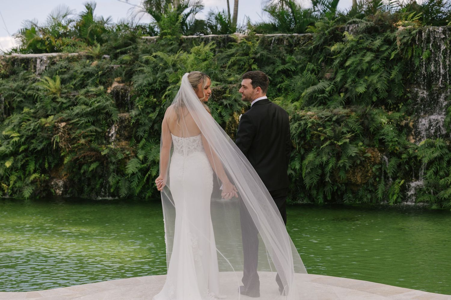 A couple shares a romantic moment on a dock by a pond with lush tropical greenery in the background.