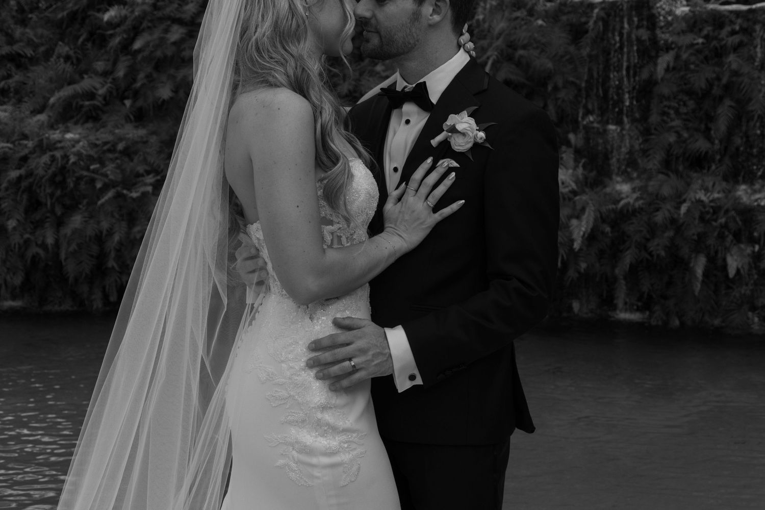 A romantic black and white wedding photo of a couple embracing by a lake, with the bride wearing a flowing veil.