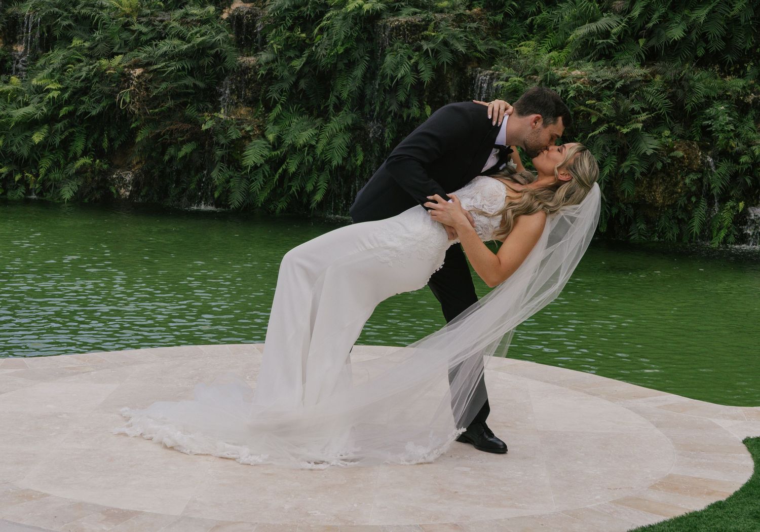 A romantic couple in wedding attire share an intimate moment while posing near a serene green lake.