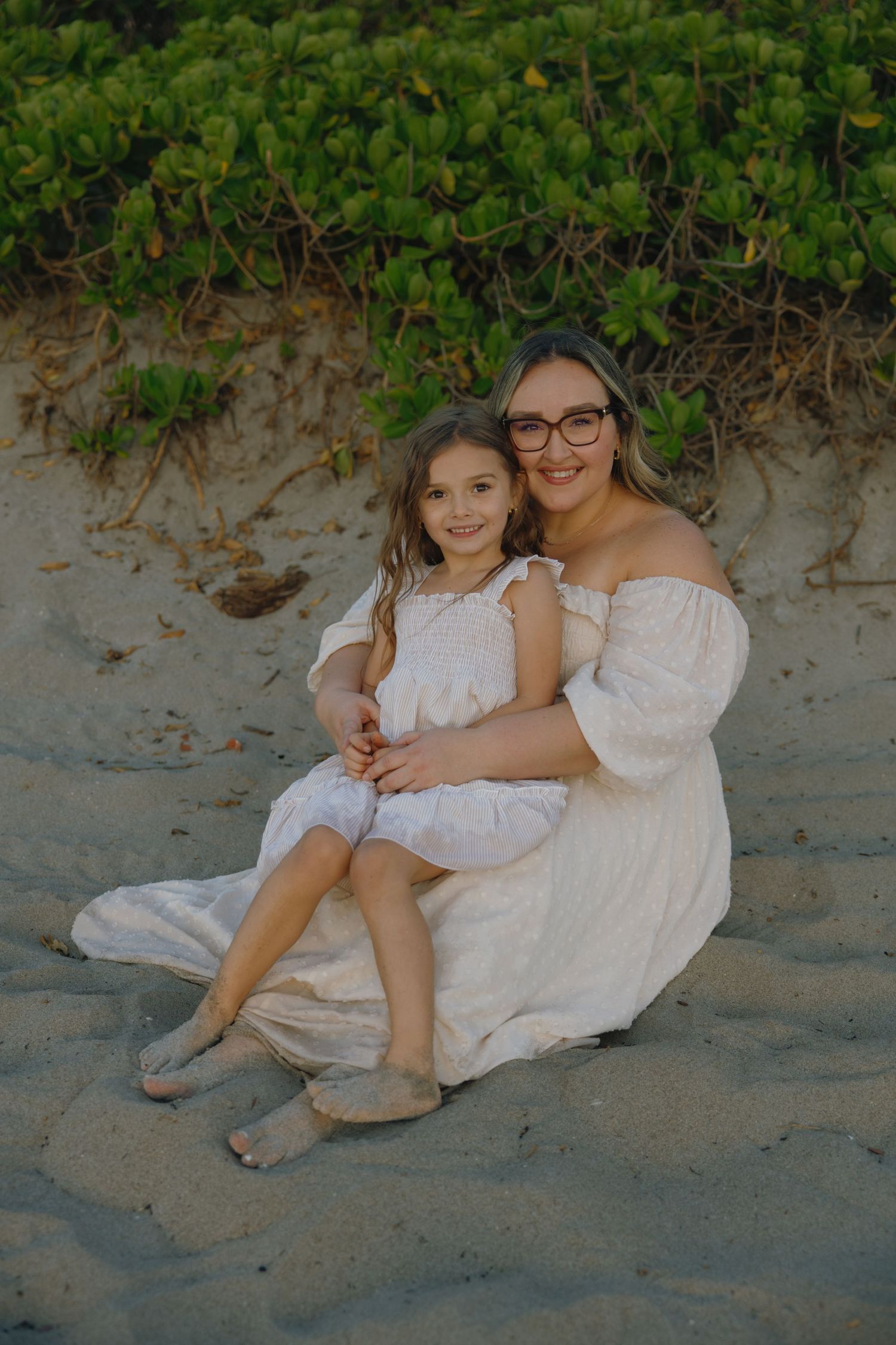 Two people in white dresses sit together on the beach near green foliage.