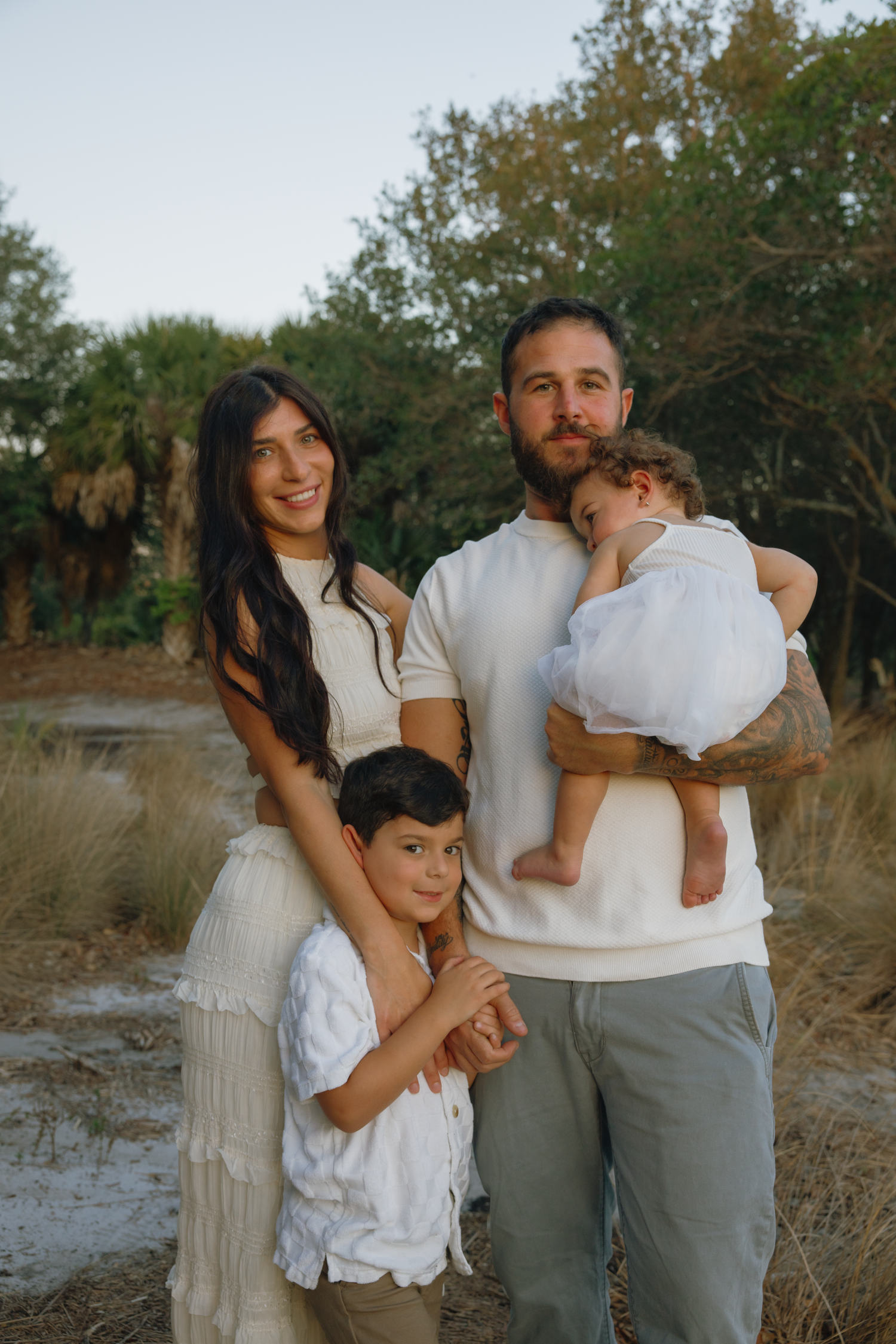 Family dressed in white and neutral tones embraces warmly in natural outdoor setting.