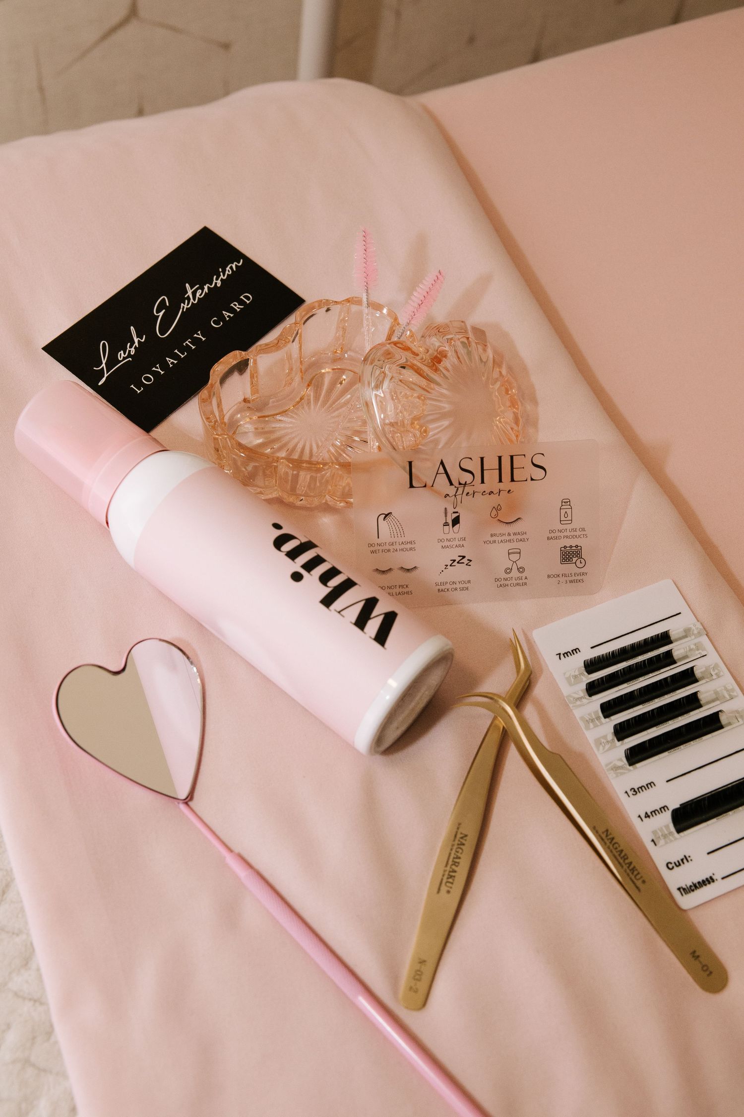 Beauty supplies and lash treatment tools arranged on pink surface with gold accents.