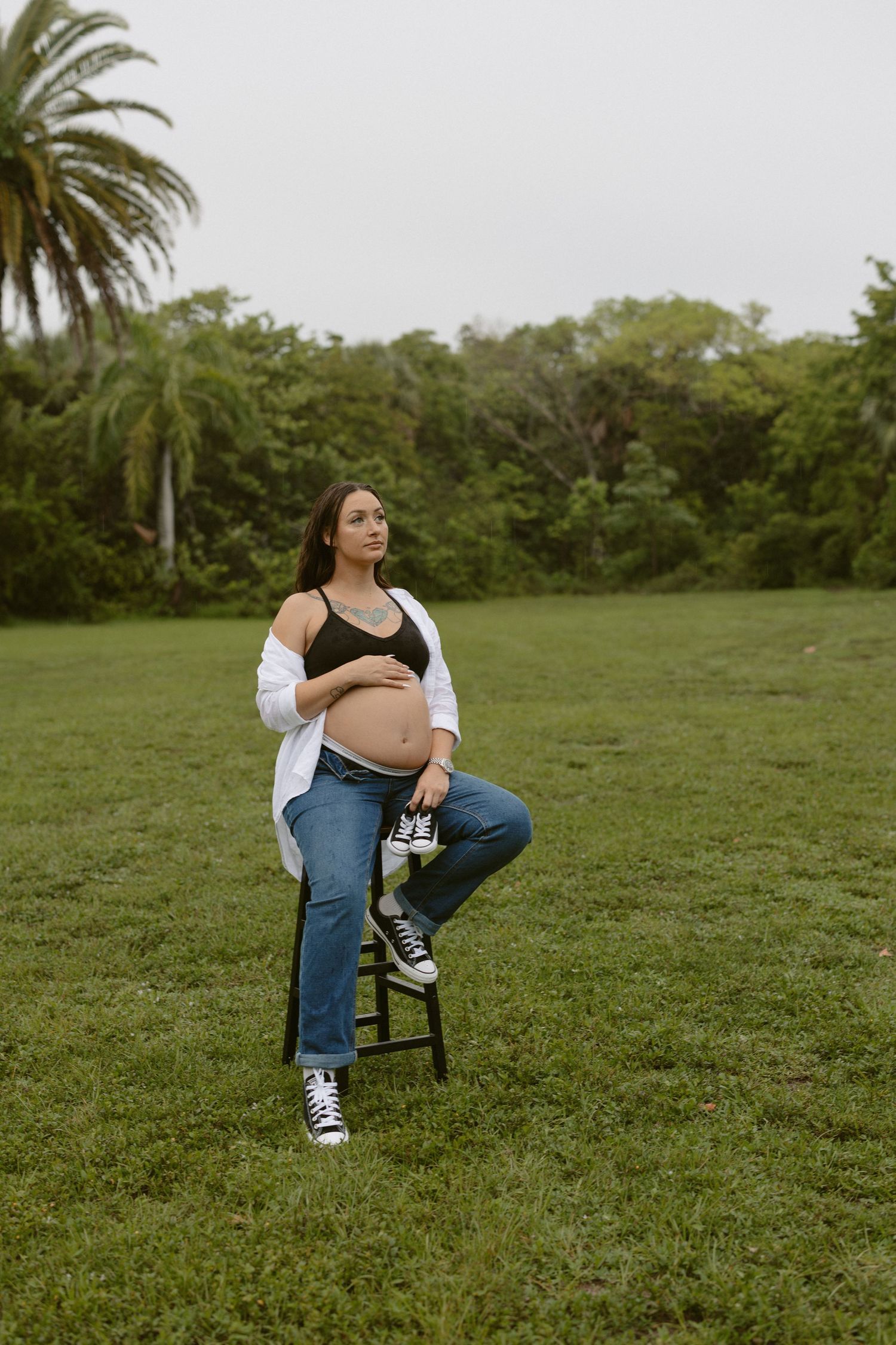 A person in jeans and a black top sits casually on a black chair in a grassy field with palm trees in the background.