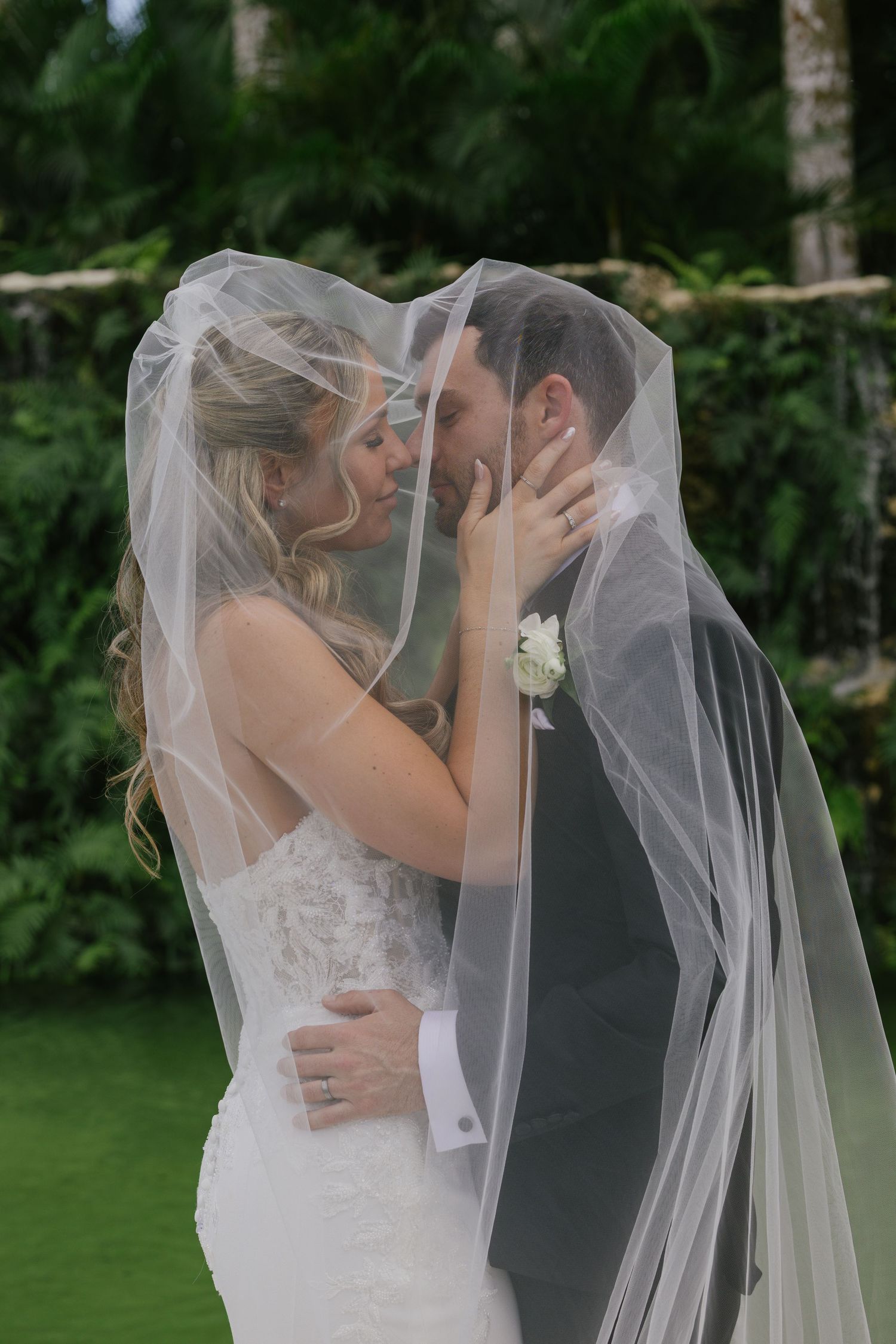 A romantic wedding couple shares an intimate kiss under a flowing veil in a lush garden setting.