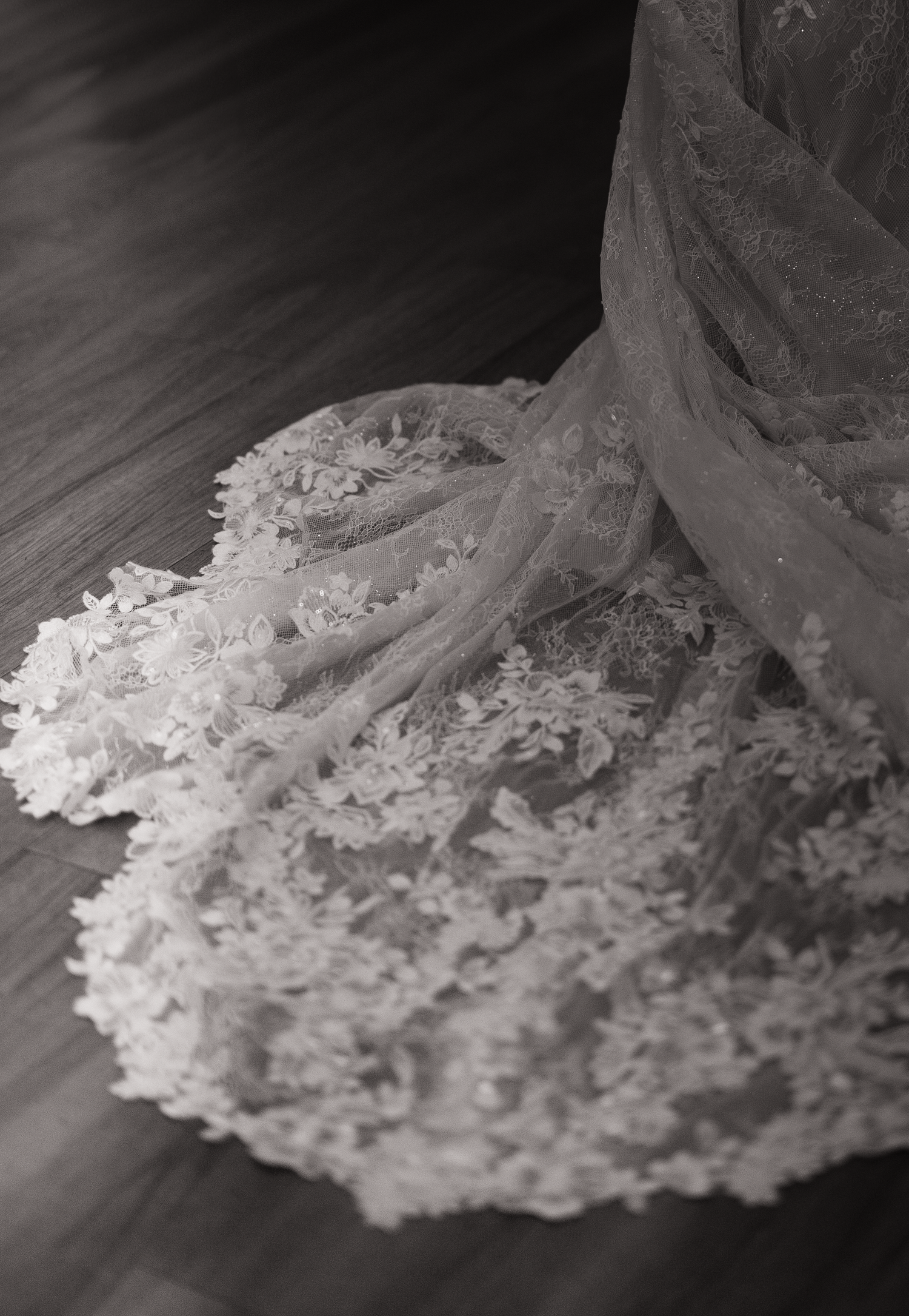 Close-up black and white photo of delicate lace wedding dress fabric trailing on dark wooden floor.