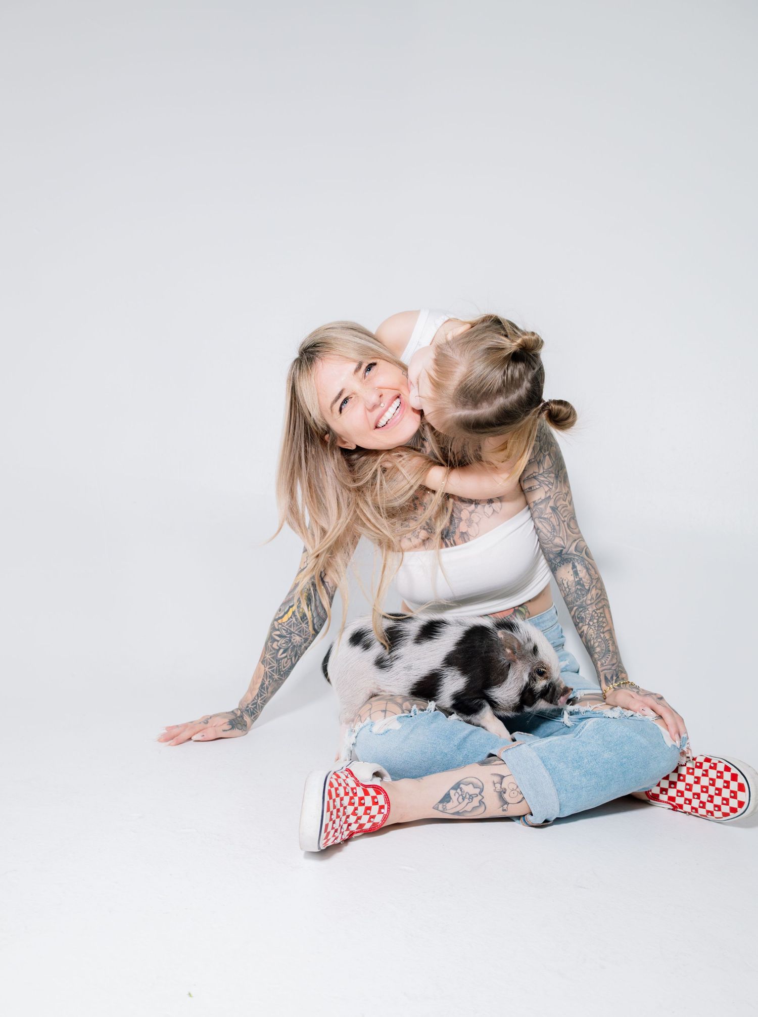 Tattooed woman sitting cross-legged on white background, smiling as a young child hugs her from behind.