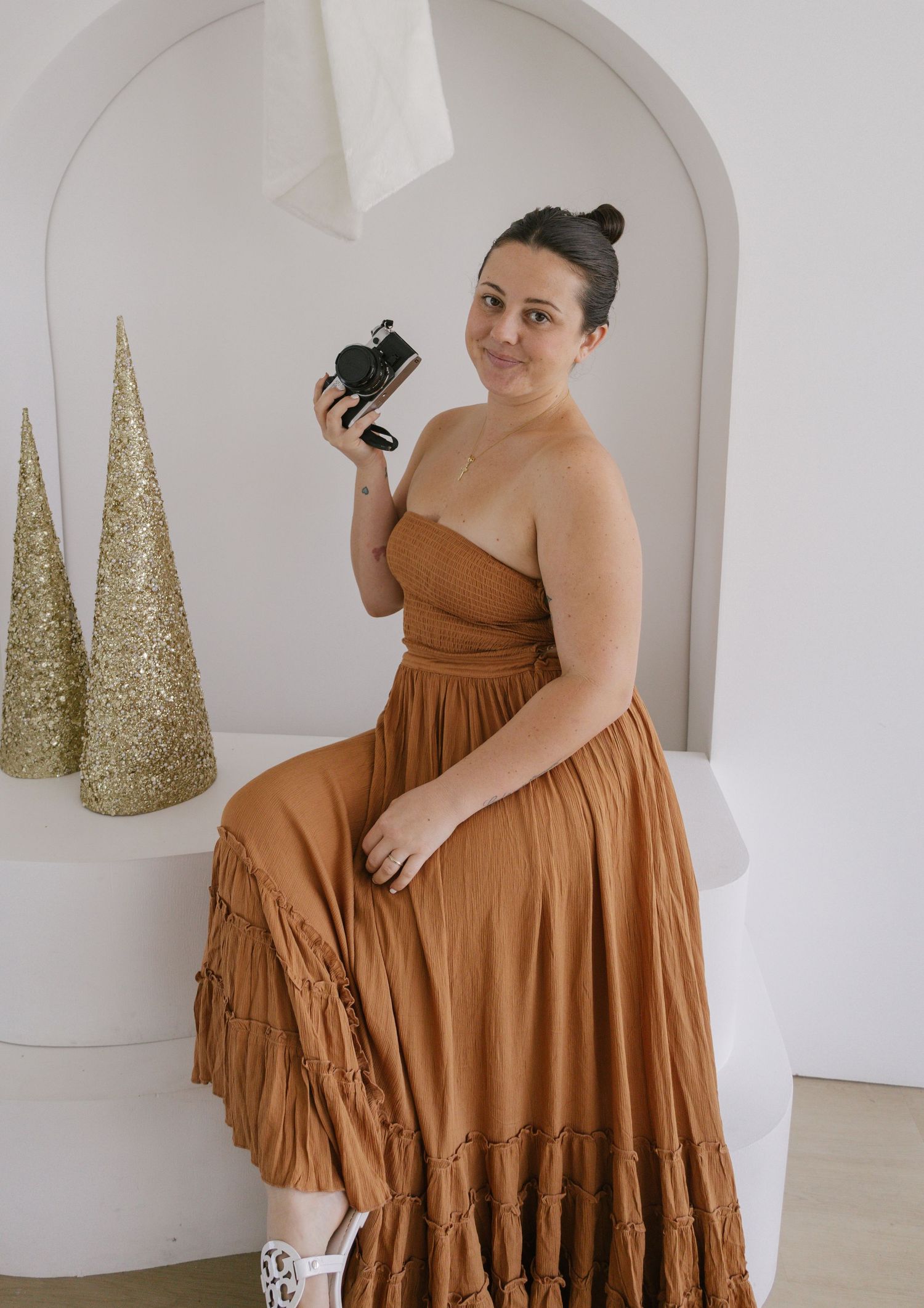 Woman in rust-colored strapless maxi dress holding a camera, seated near gold Christmas trees in a white studio.