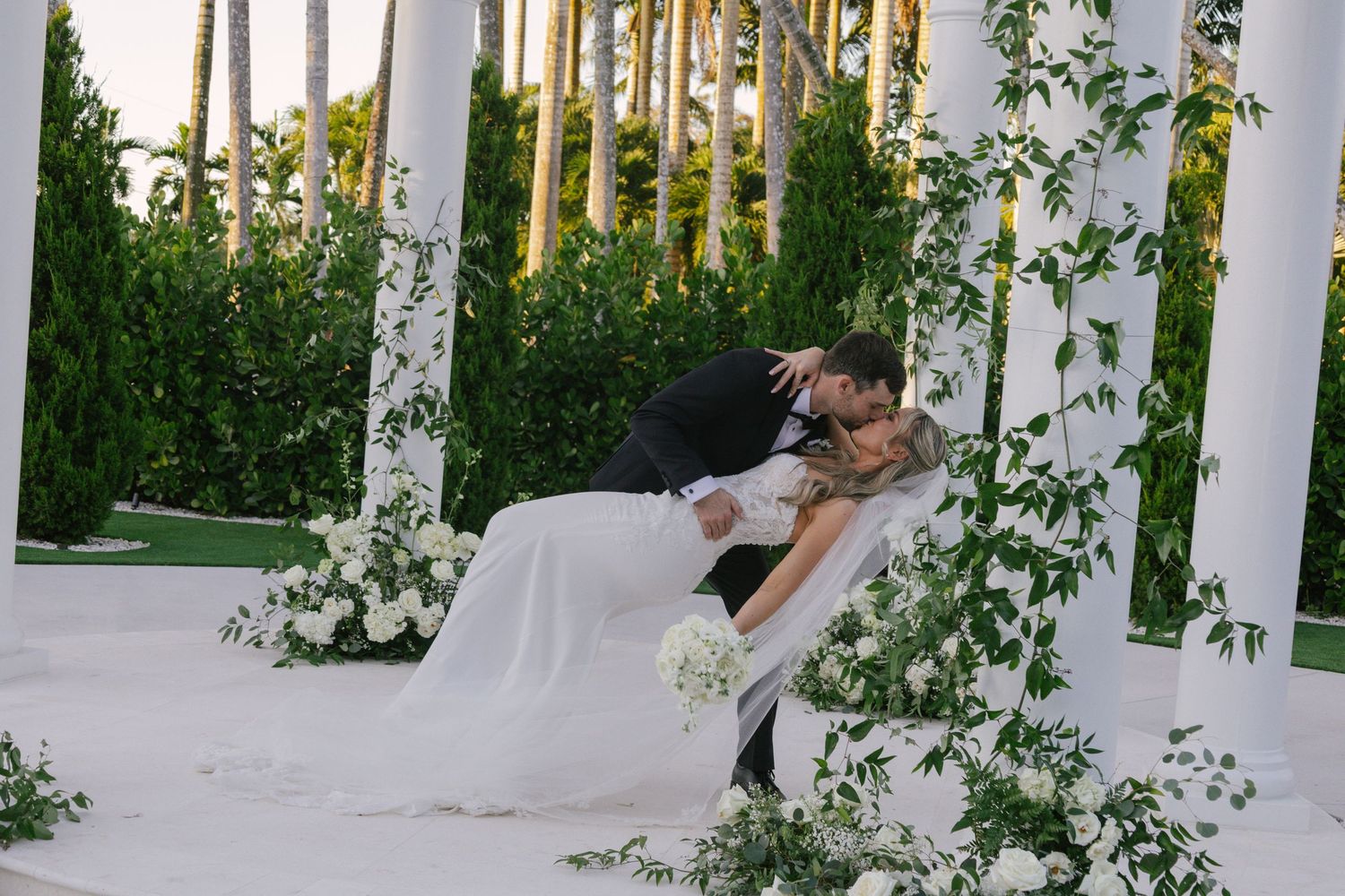 A couple shares a romantic dip kiss under white pillars surrounded by lush greenery and floral decor at an outdoor venue.