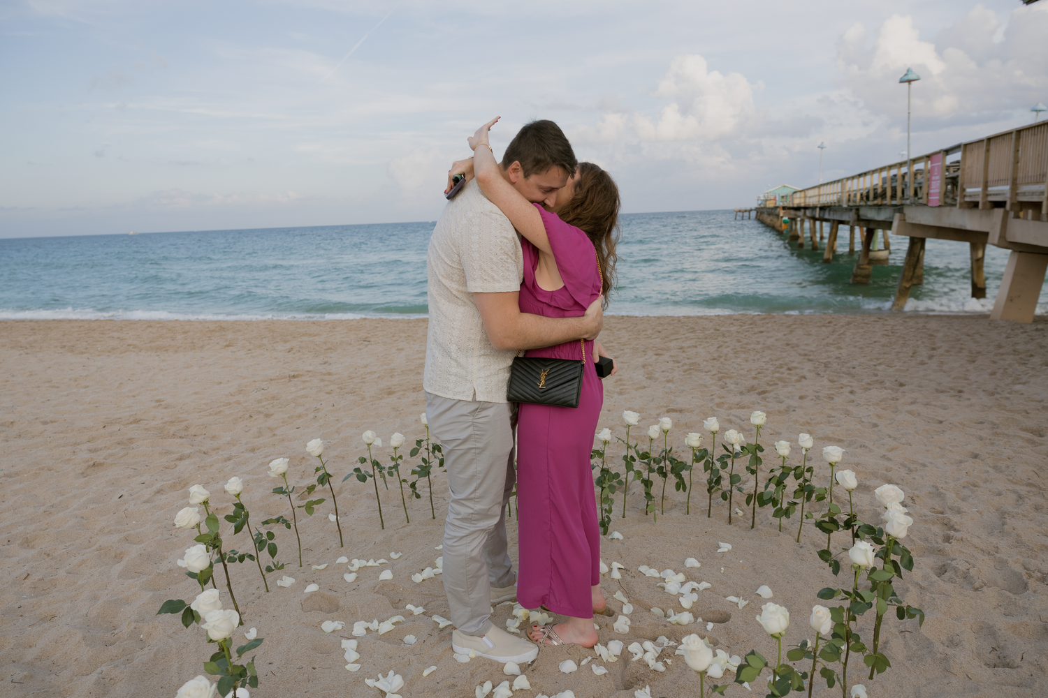 A romantic beach proposal setup with white flower petals arranged in a heart shape on the sand at sunset.