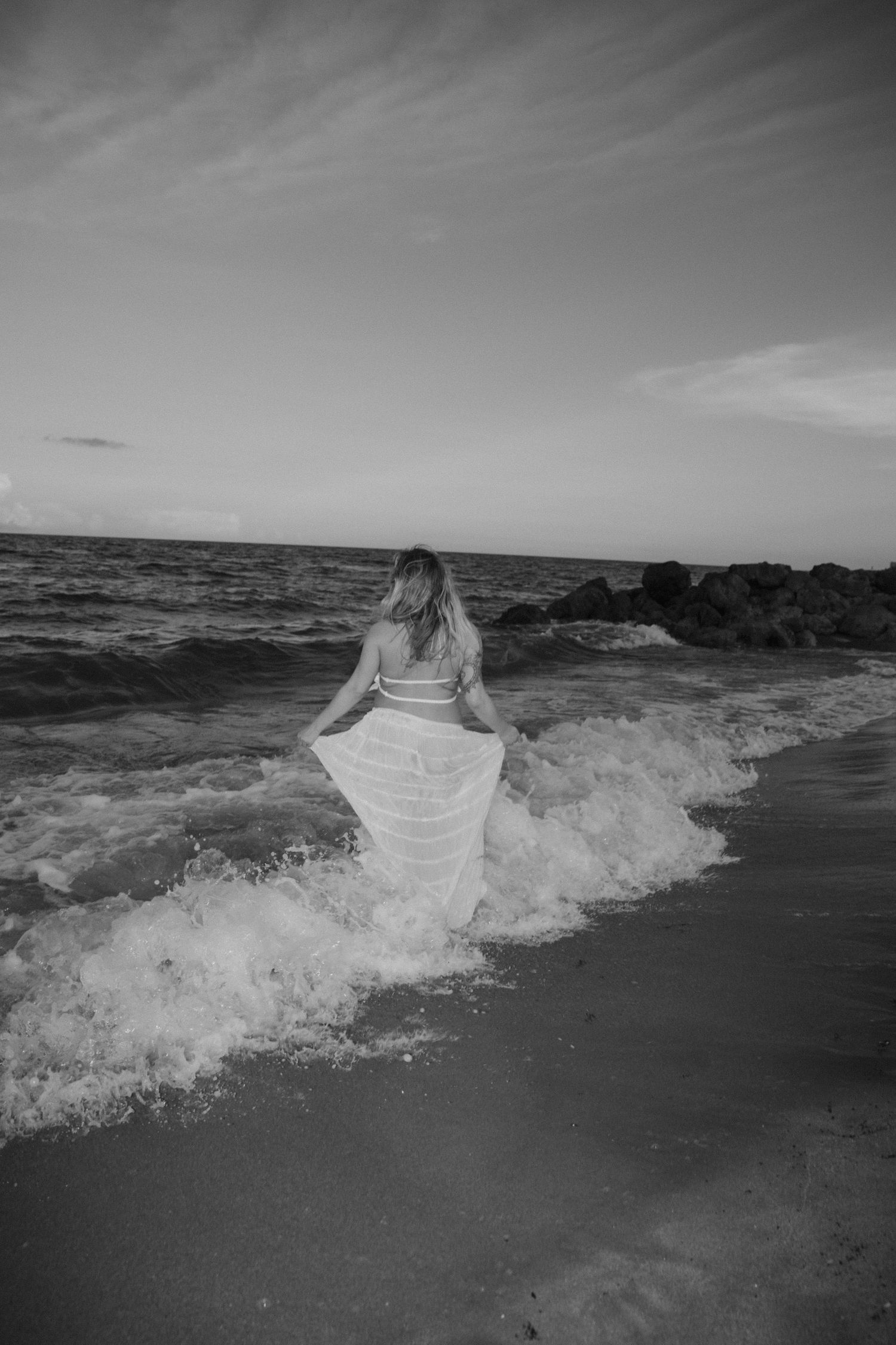 Monochrome image of woman in white dress walking through ocean waves at dusk.