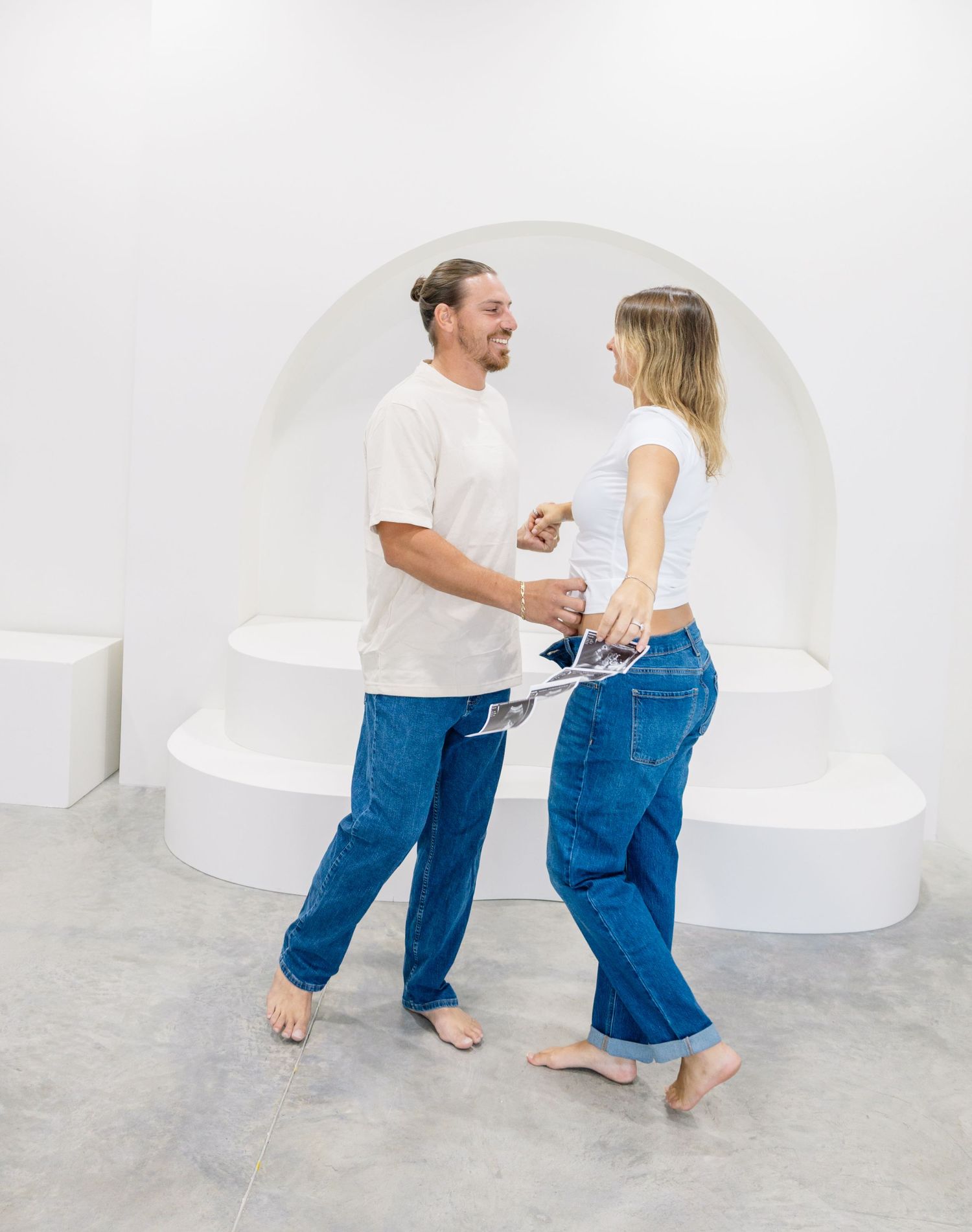 A man and woman in matching white tops and blue jeans dance barefoot in a minimalist white gallery space.