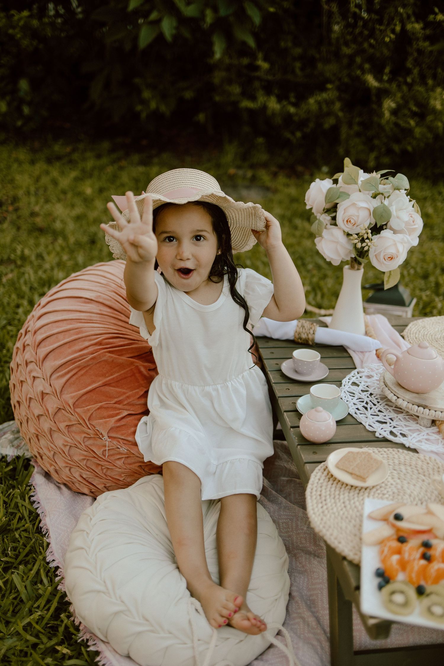 A child in a white dress and sun hat sits at an outdoor tea party setting with pink decor and floral arrangements.