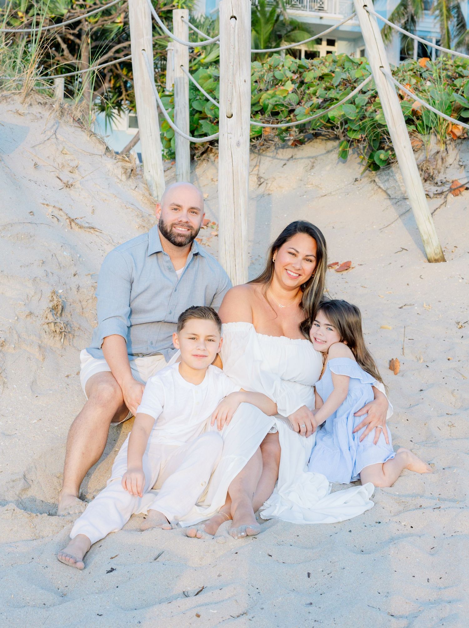 Family sitting together on a sandy beach slope with greenery and dunes in the background.