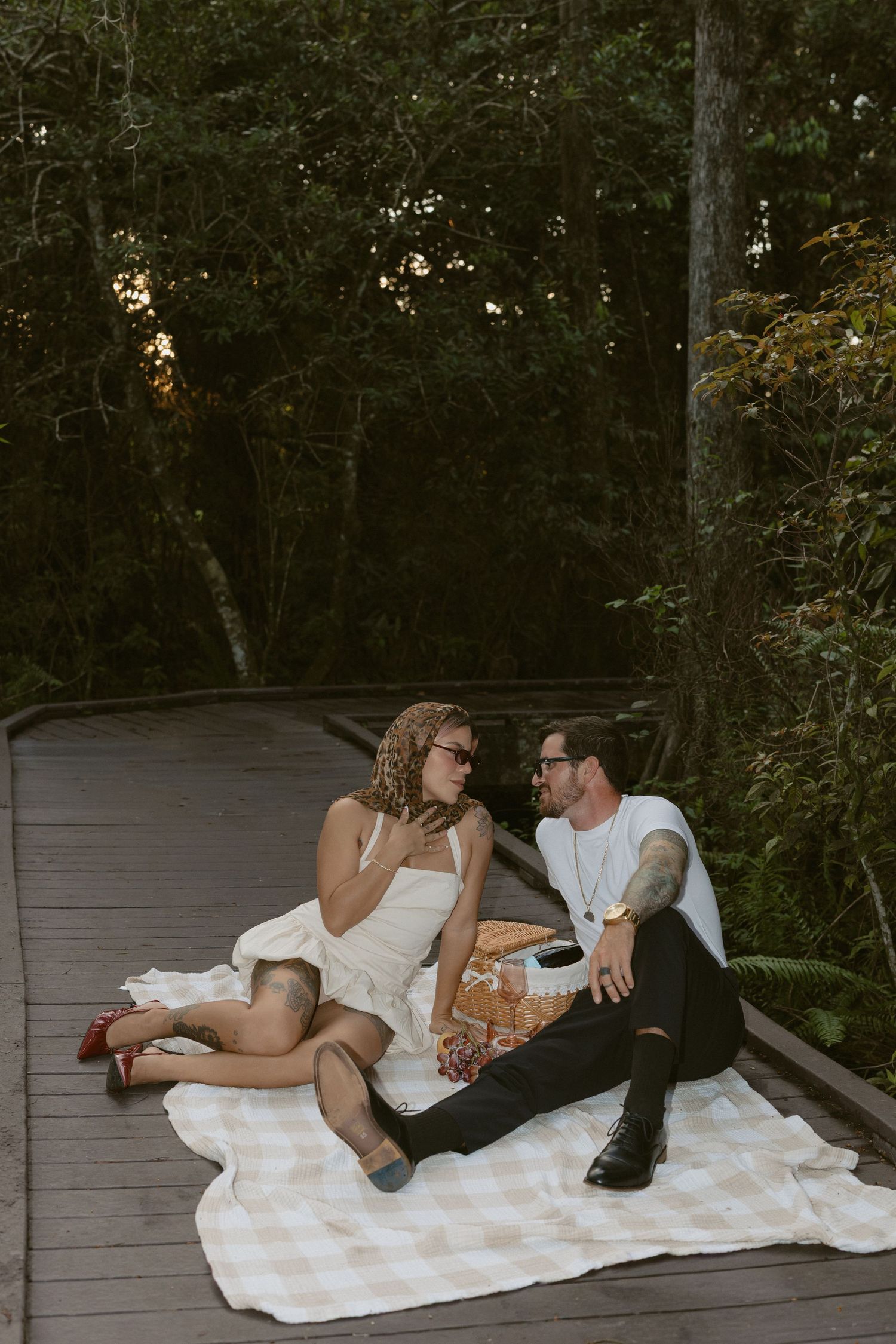 A romantic couple shares intimate moments on a blanket laid out on a wooden deck surrounded by trees at dusk.