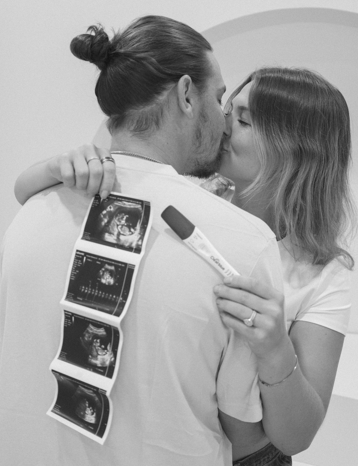 Couple kissing while holding ultrasound photos and pregnancy test, announcing their pregnancy in black and white photo.