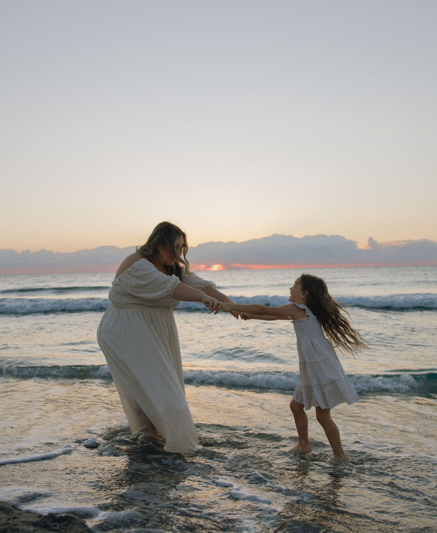 Two people in white dresses dance playfully in the ocean waves at sunset.
