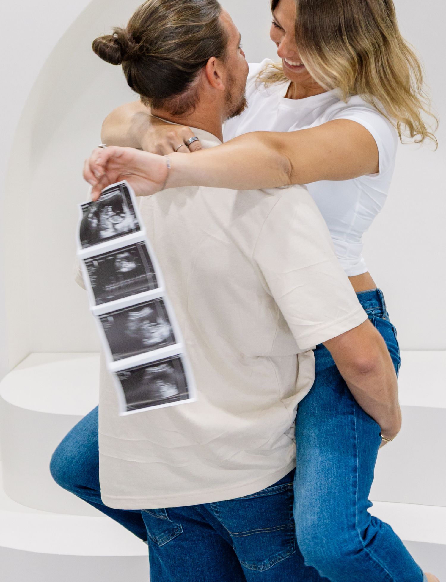 Couple embracing and holding ultrasound photos, announcing pregnancy in a bright white studio setting.