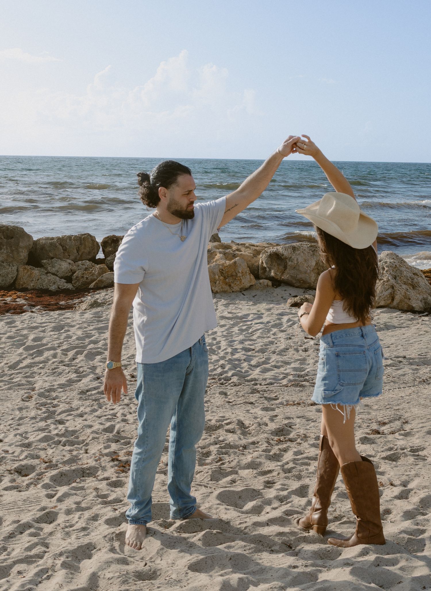 Couple dancing on a sandy beach near rocky shore, woman in cowboy hat and boots, man in white tee and jeans.