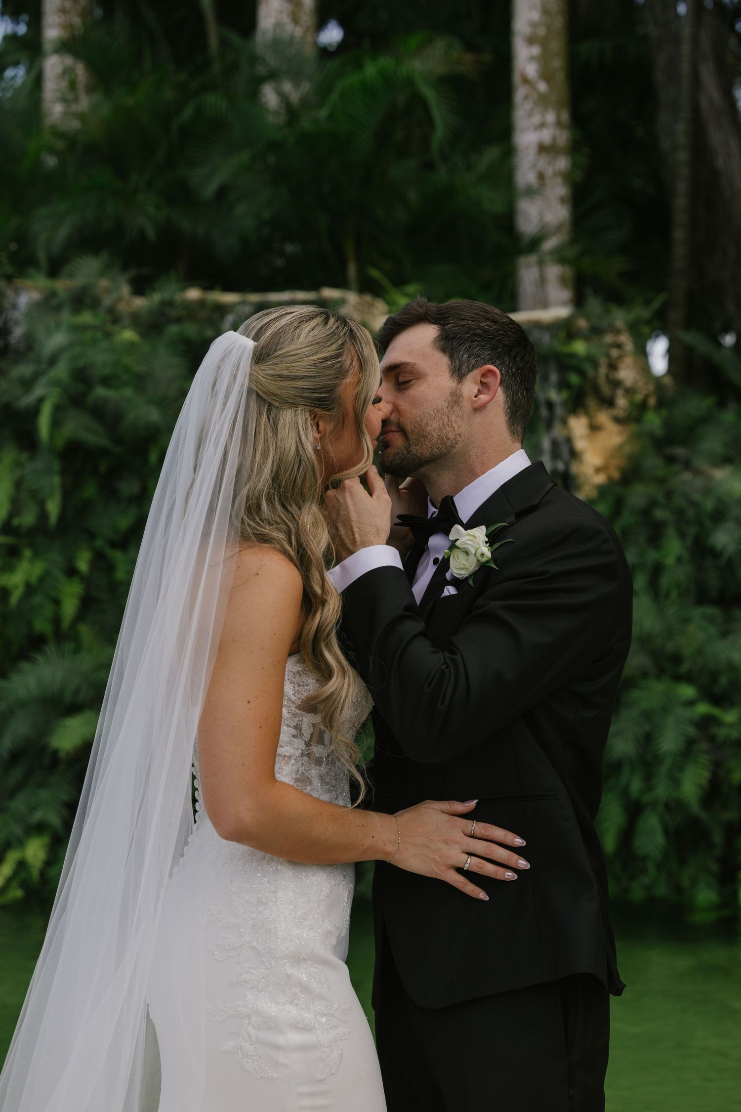 A bride and groom share romantic kisses in a lush garden setting with trees in the background.