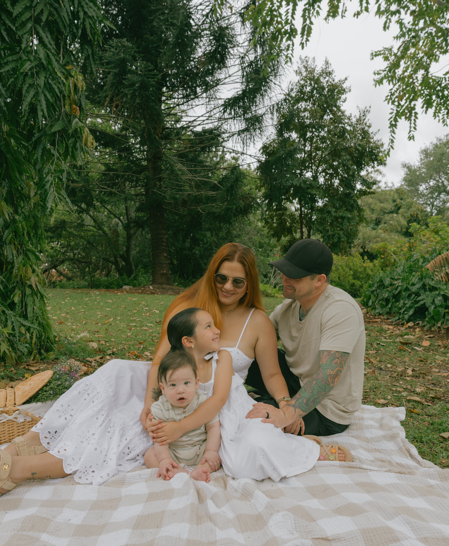 Family enjoying a peaceful picnic under willow trees on a white blanket in a lush garden setting.
