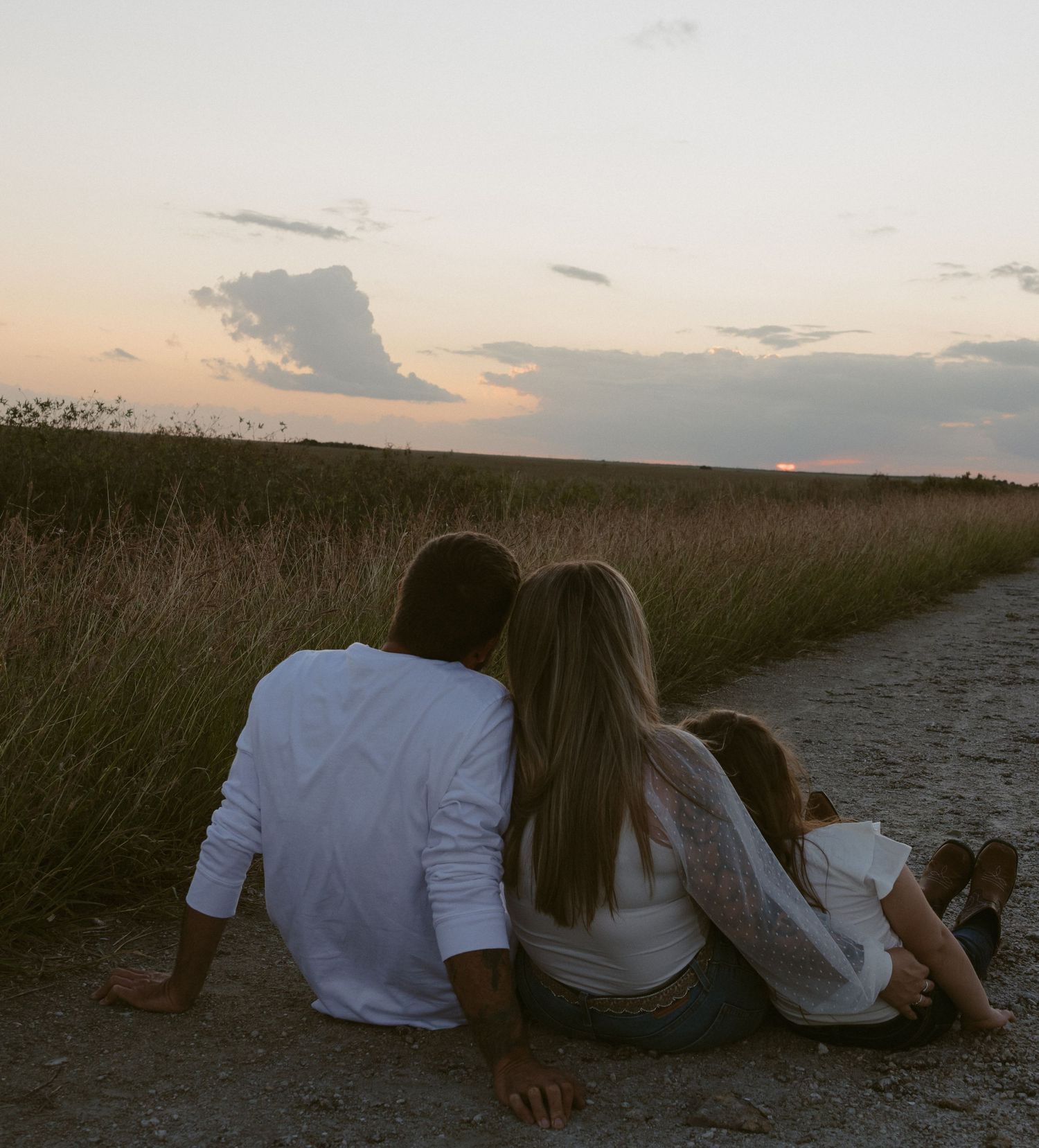 A family sits together watching a peaceful sunset over tall grass fields along a dirt path.