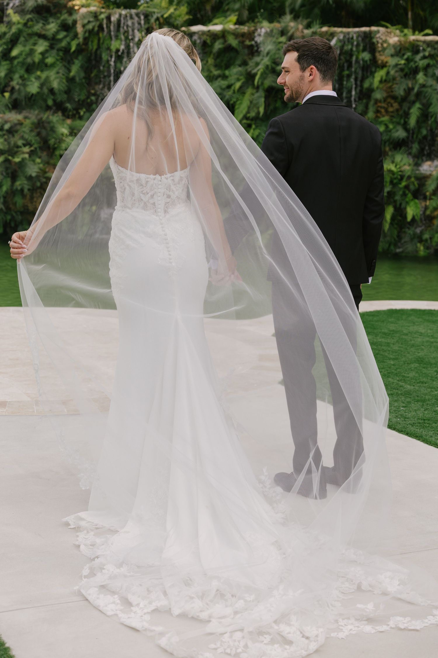 A flowing cathedral-length wedding veil with delicate lace trim trails behind a white bridal gown in an outdoor garden setting.