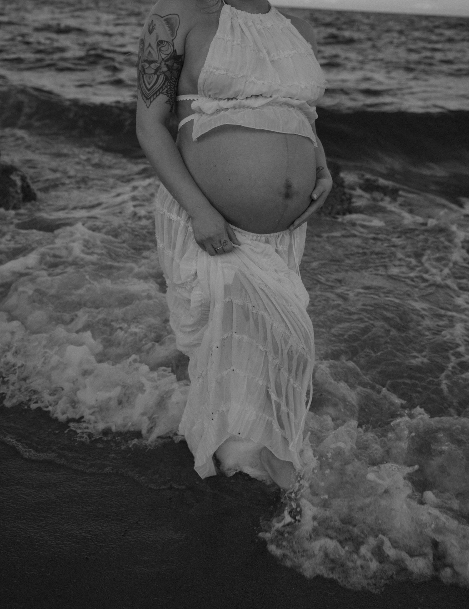 Black and white photo highlighting pregnant belly against oceanic background with waves.