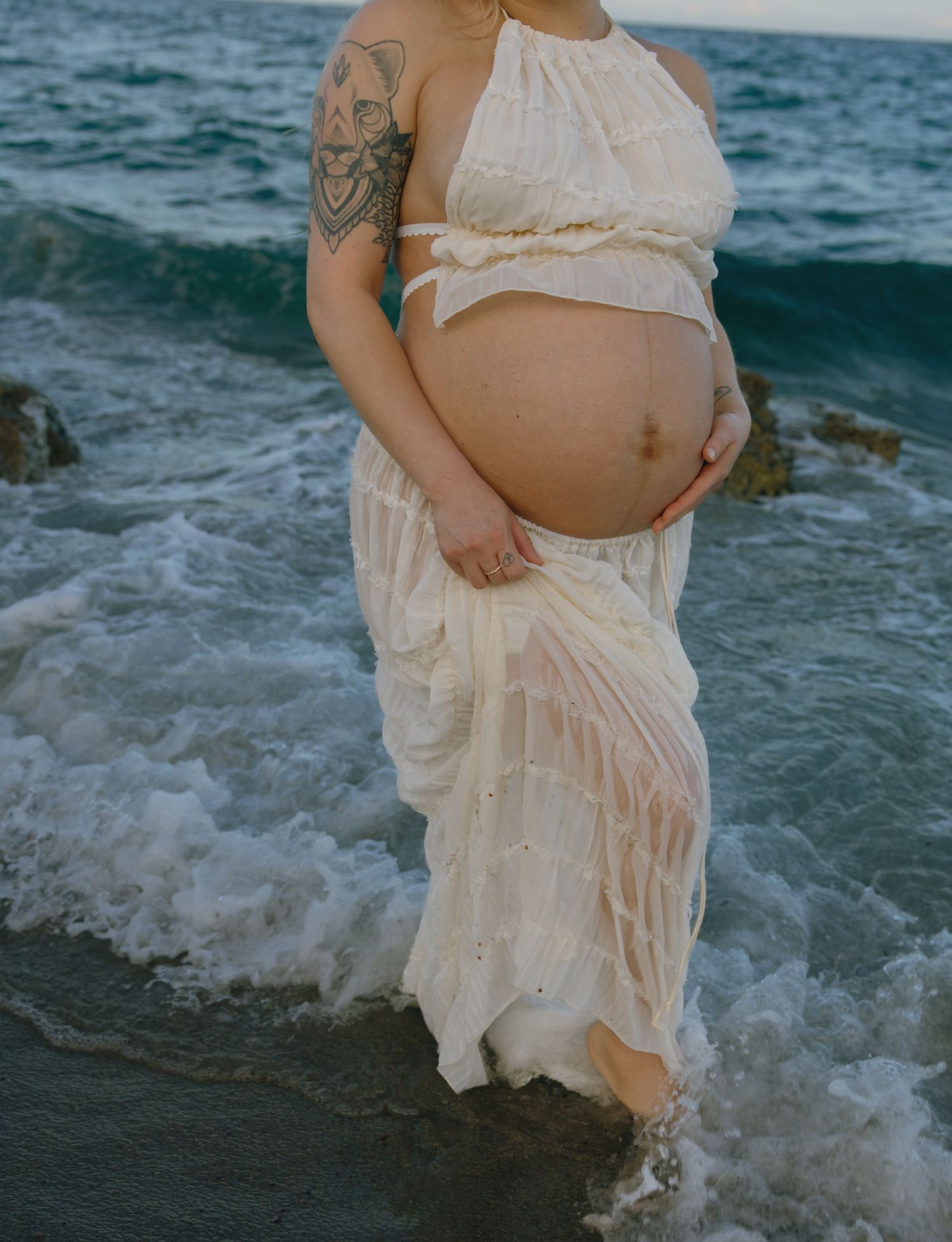 A pregnant person in a flowing white dress stands at the ocean's edge with visible arm tattoos and waves lapping at their feet.