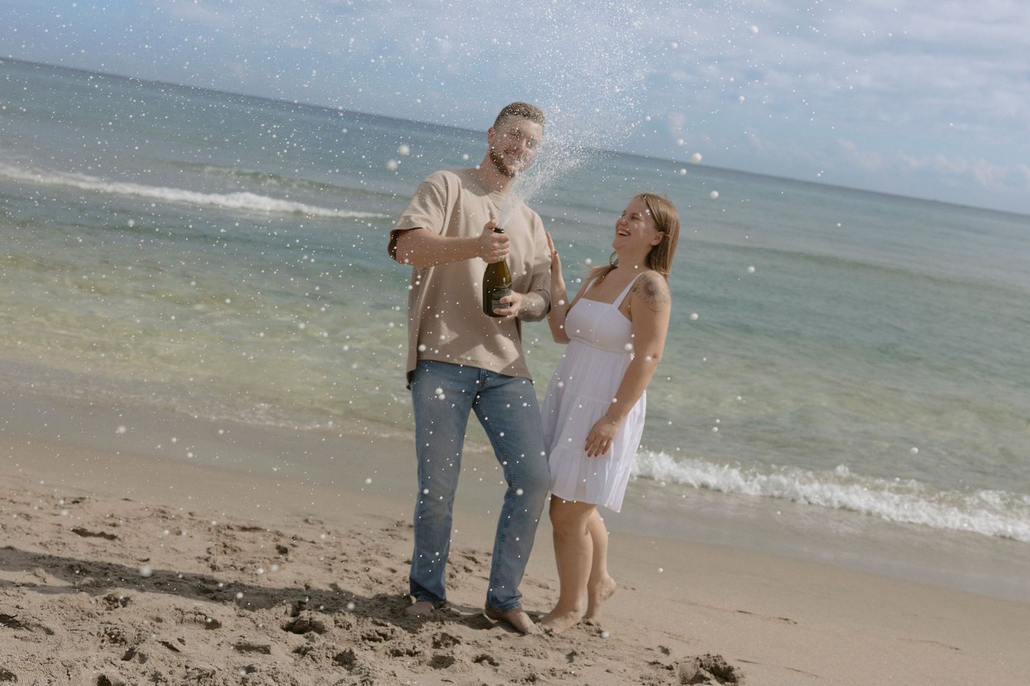 A couple shares a joyful moment together on a sunny beach with waves crashing in the background.