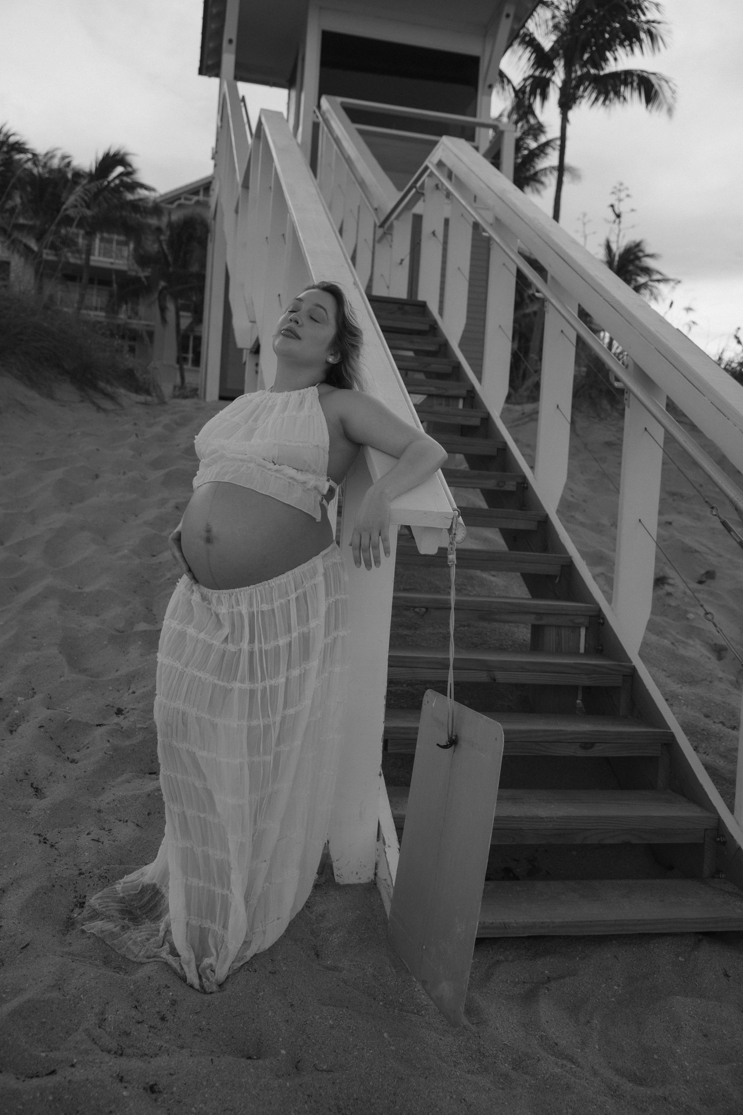 Woman in sheer white dress poses near beach stairs and palm trees.