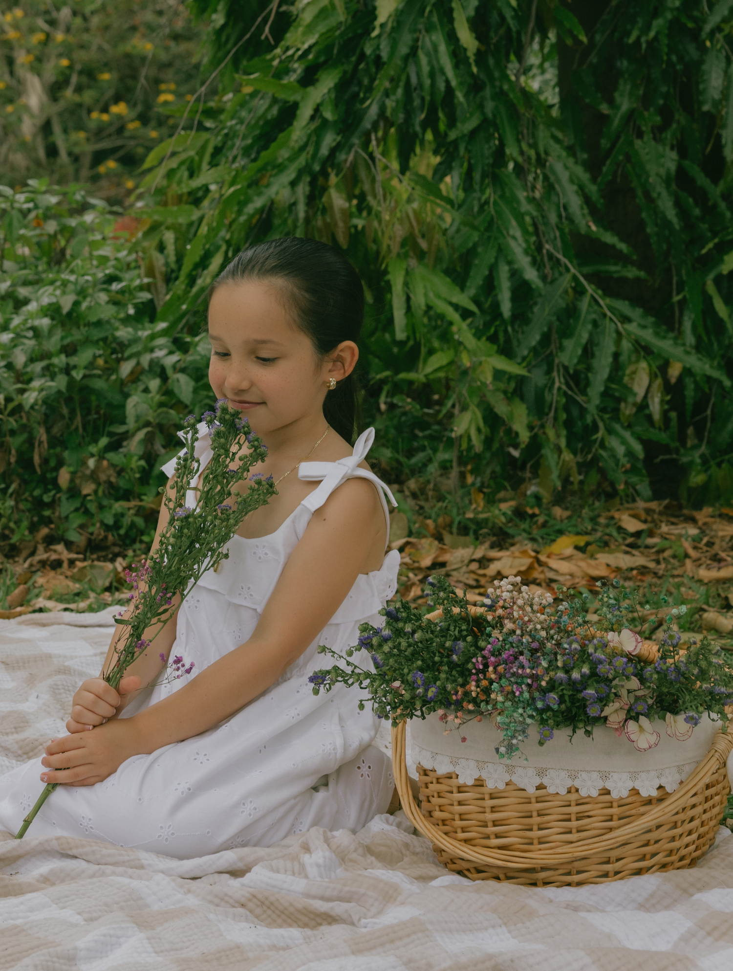 Young girl in white dress holding wildflowers next to wicker basket on picnic blanket.