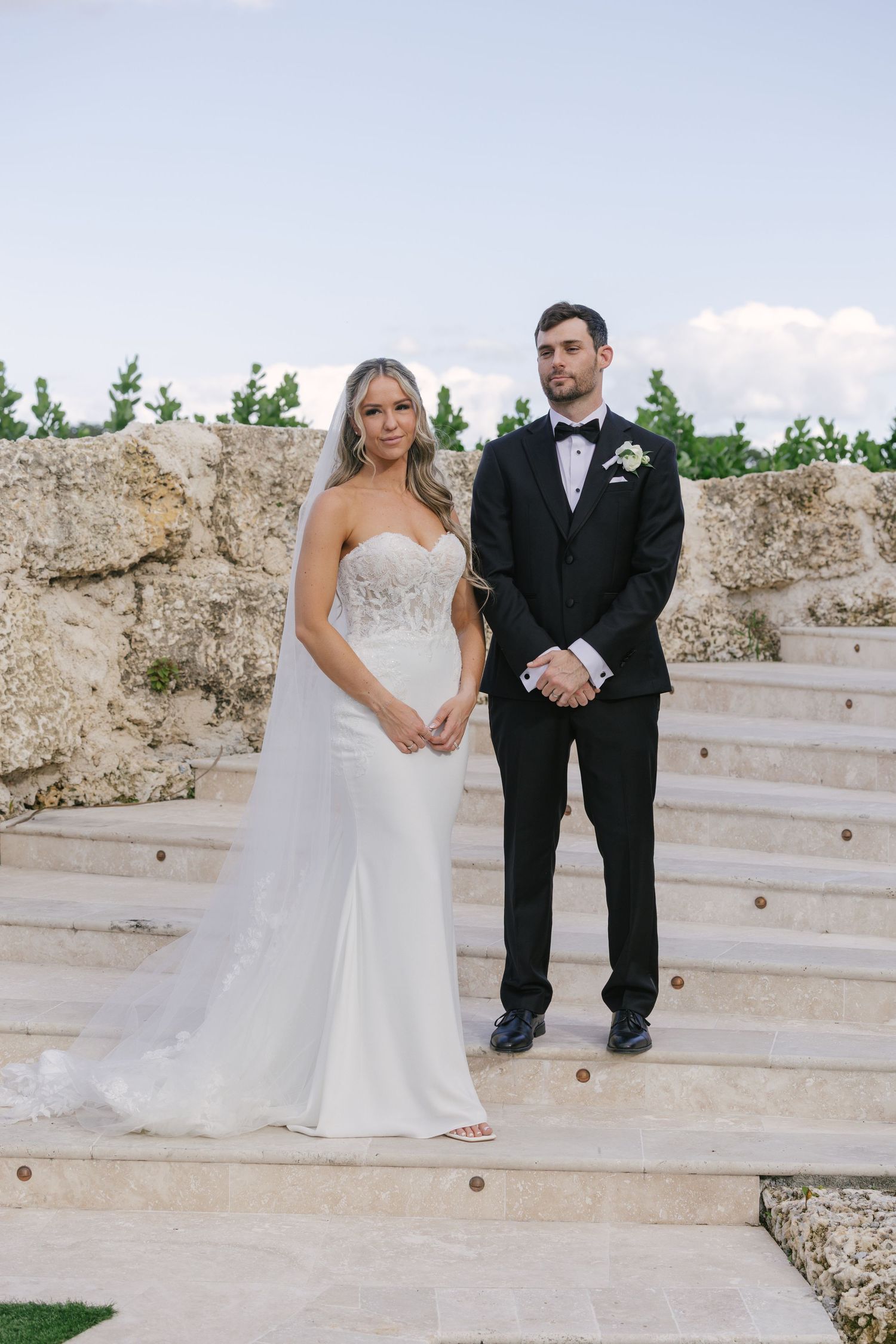 A couple in wedding attire sits together on white stone steps in an outdoor setting.