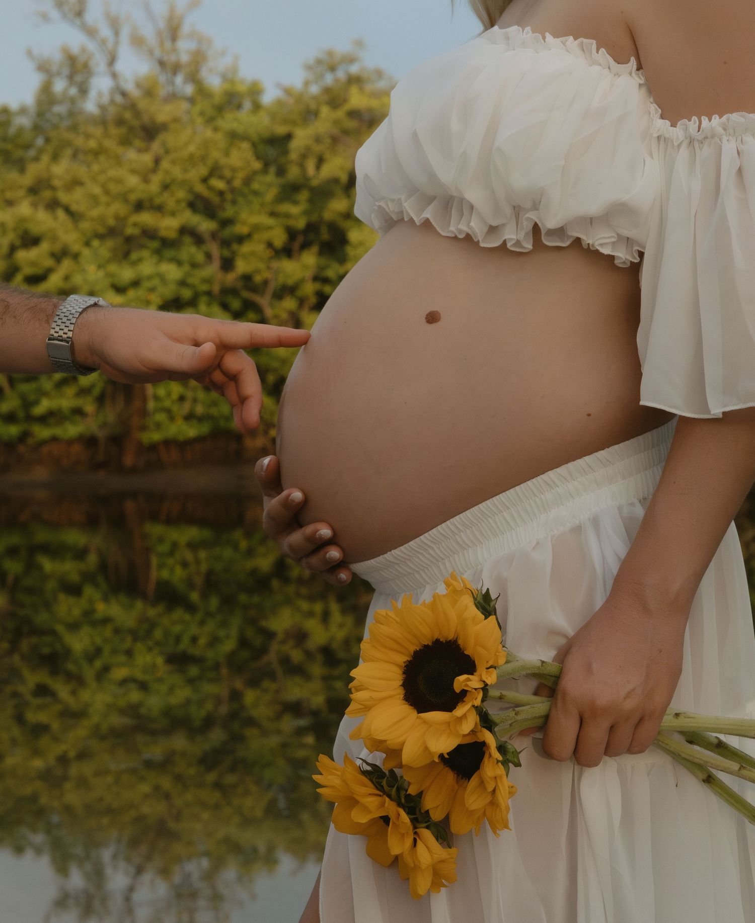 A pregnant person in a white dress holds sunflowers while standing near a pond on a sunny day.