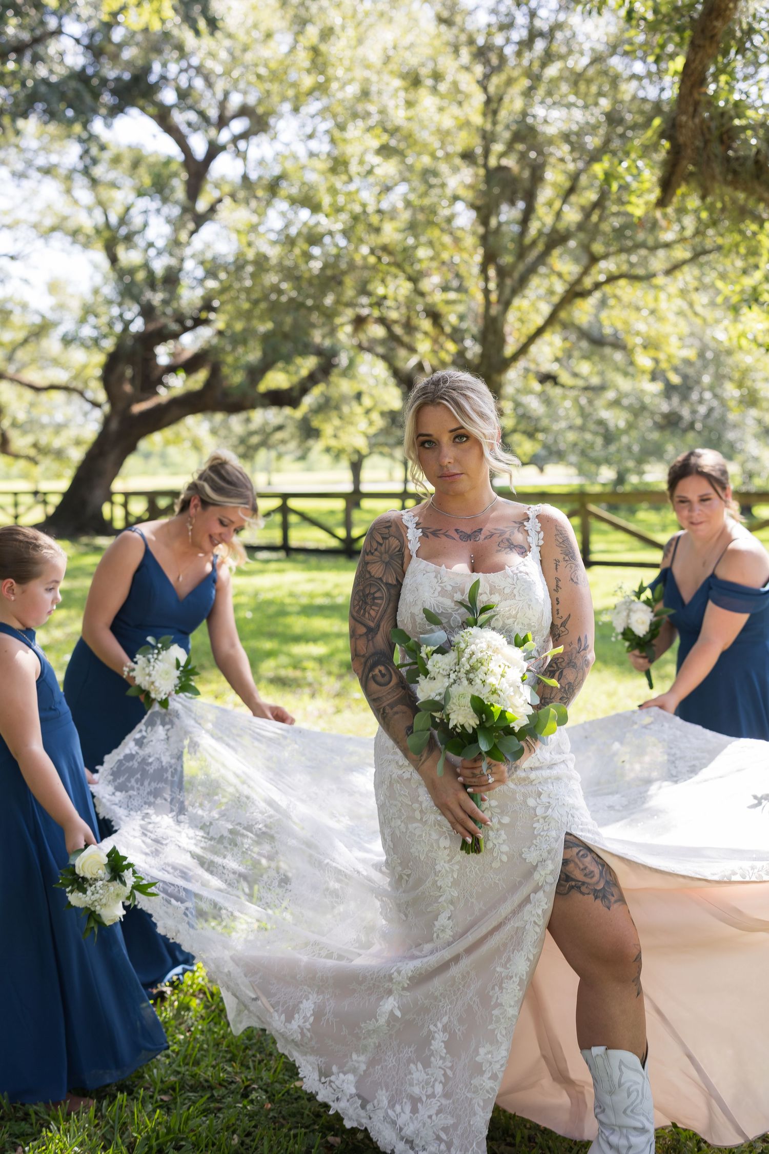 Bridesmaids in navy dresses help arrange a flowing white lace wedding dress in a sunny garden.