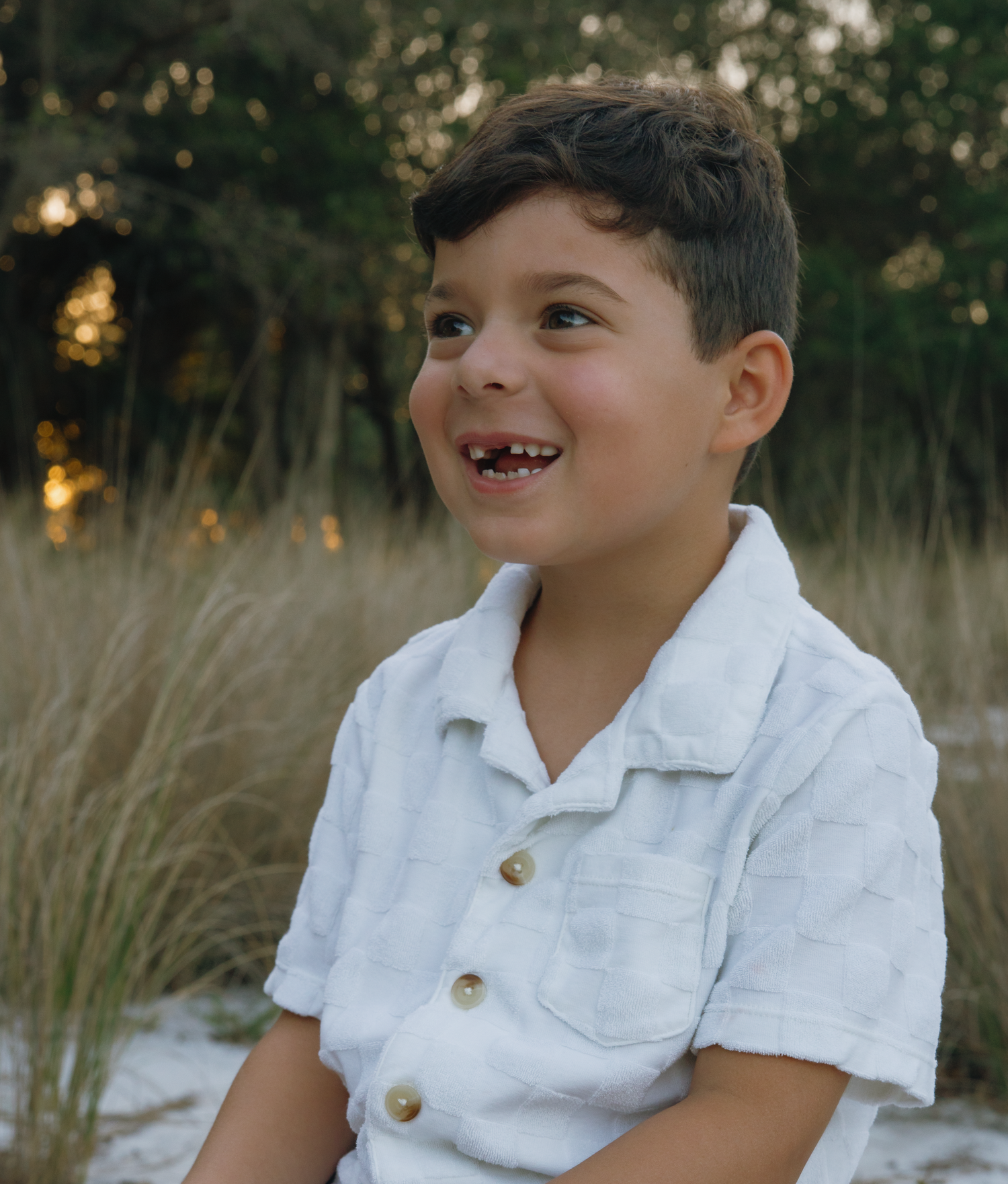 Young child in white polo shirt sits smiling at sunset on sandy beach with tall grass behind.