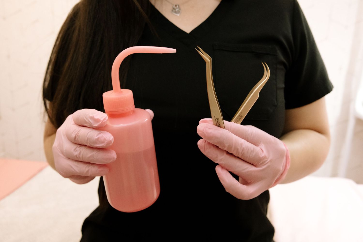 Hands holding beauty treatment tools and pink squeeze bottle against neutral background.