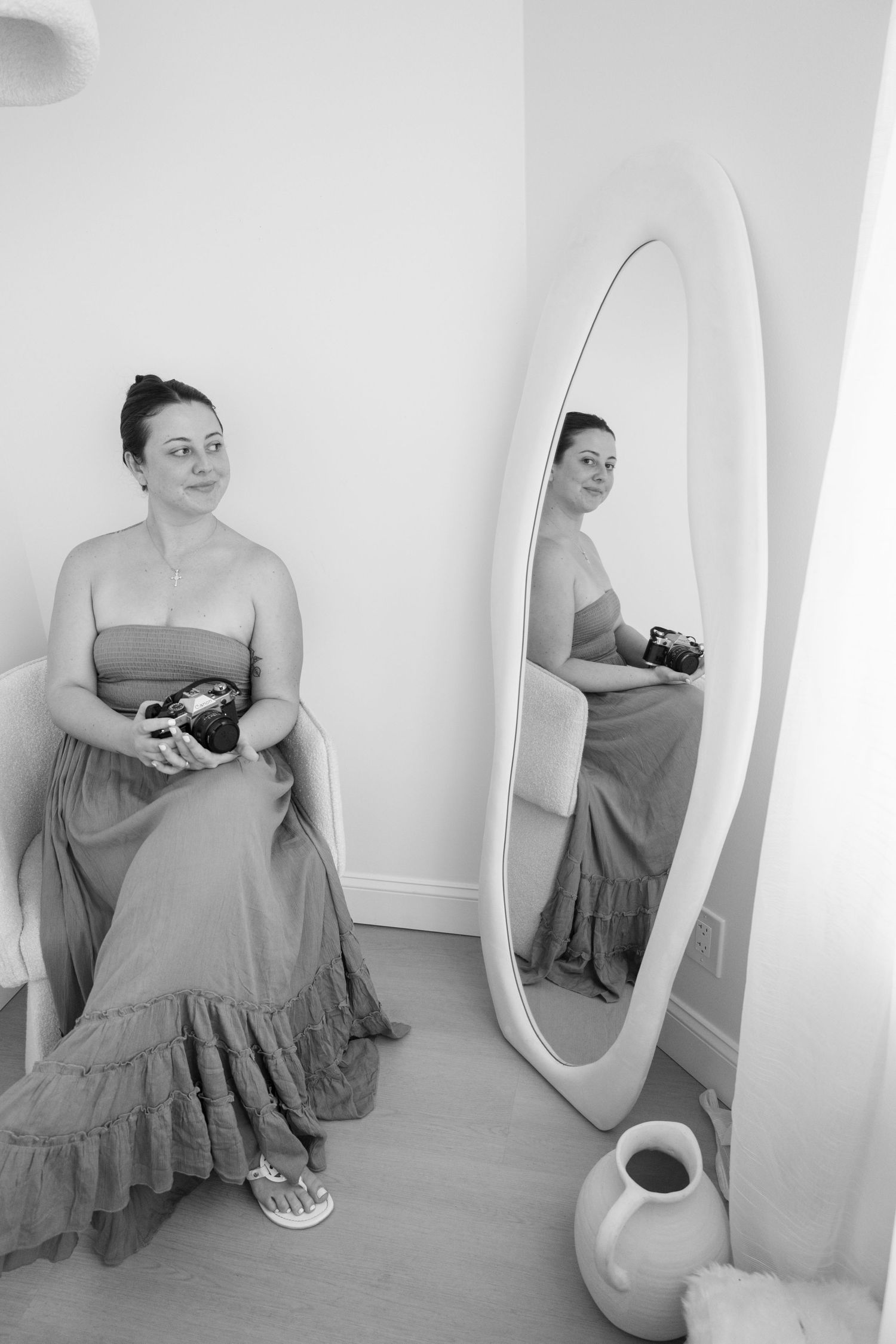 Black and white photo of a woman in a strapless maxi dress holding a camera, seated beside an oval floor mirror.