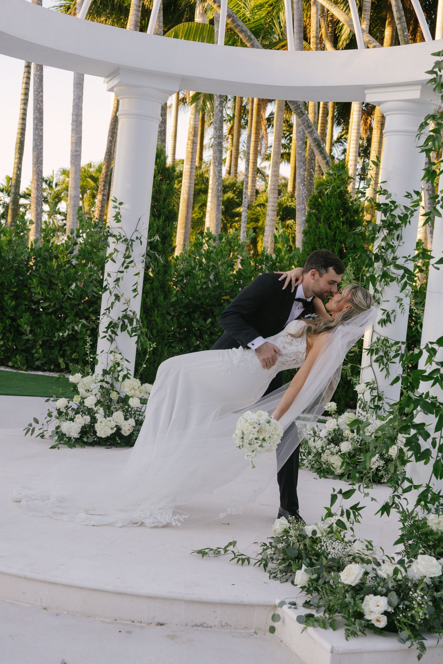 A romantic wedding couple shares an intimate dance dip moment under a white floral gazebo in a lush garden setting.