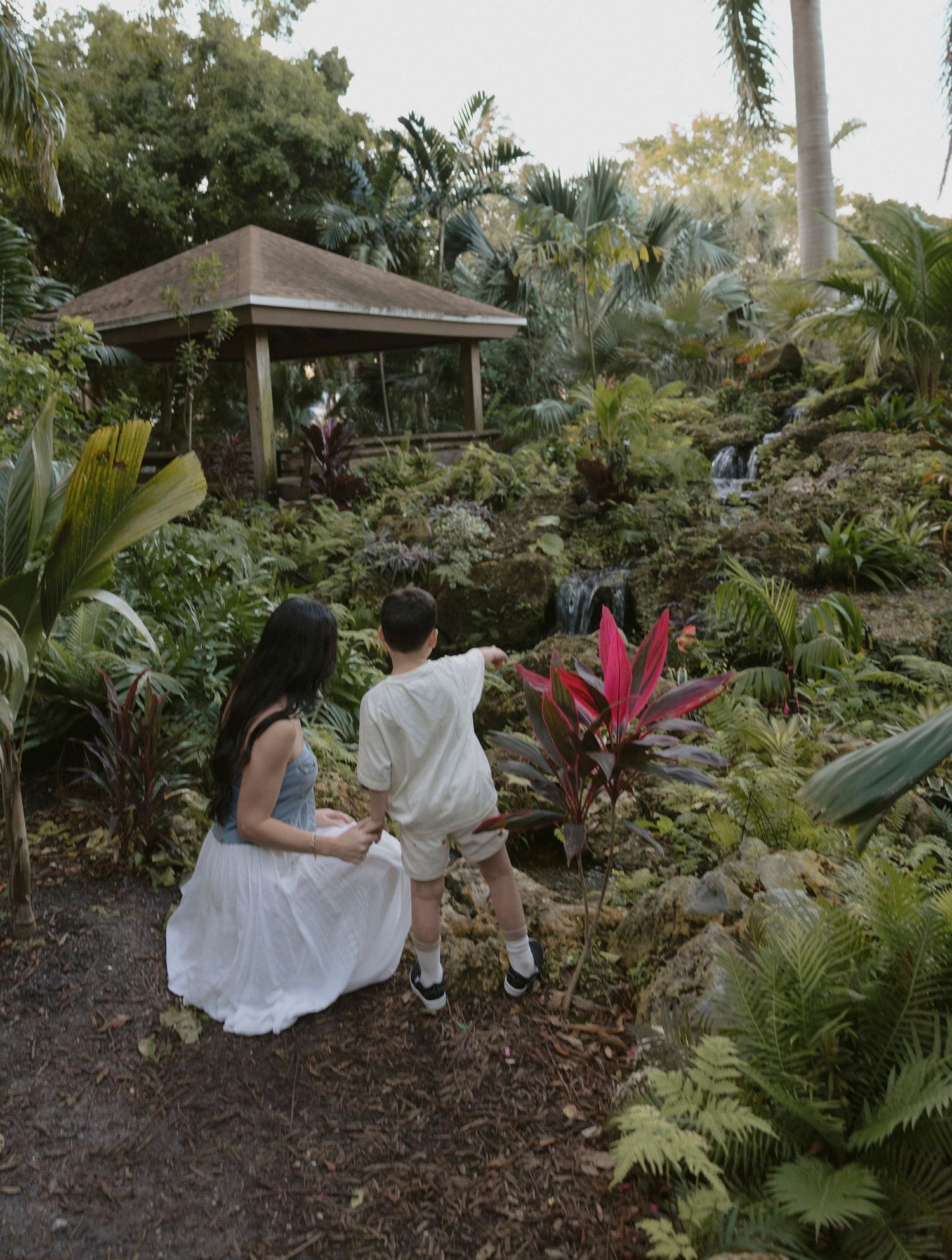 Someone in white clothing sits on the ground in a lush tropical garden setting with palm trees and ferns surrounding them.